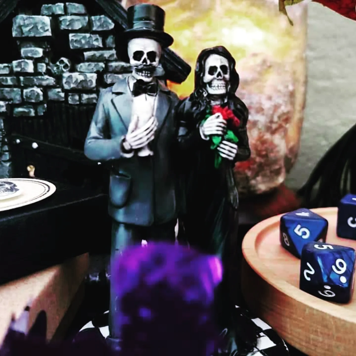 Decorative skeleton couple dressed as a bride and groom, with the skeleton groom wearing a top hat and tuxedo, and the skeleton bride in a dress holding a red rose, standing in a setting with gaming dice on a table nearby.
