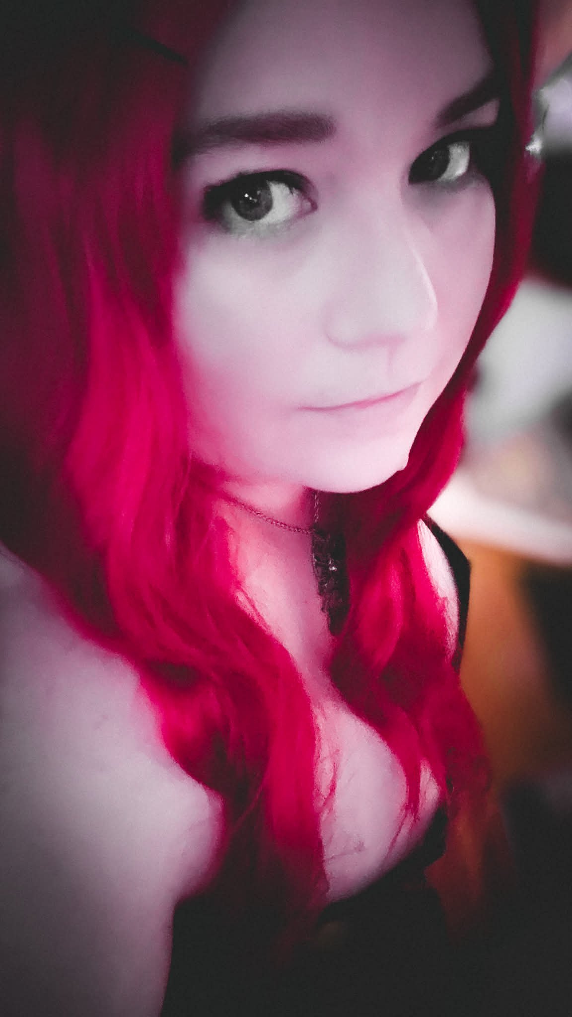 A young woman with bright red hair styled in loose waves. She has striking dark eyes and is wearing light makeup. She is looking at the camera with a slight smile, wearing a black top and a delicate necklace.