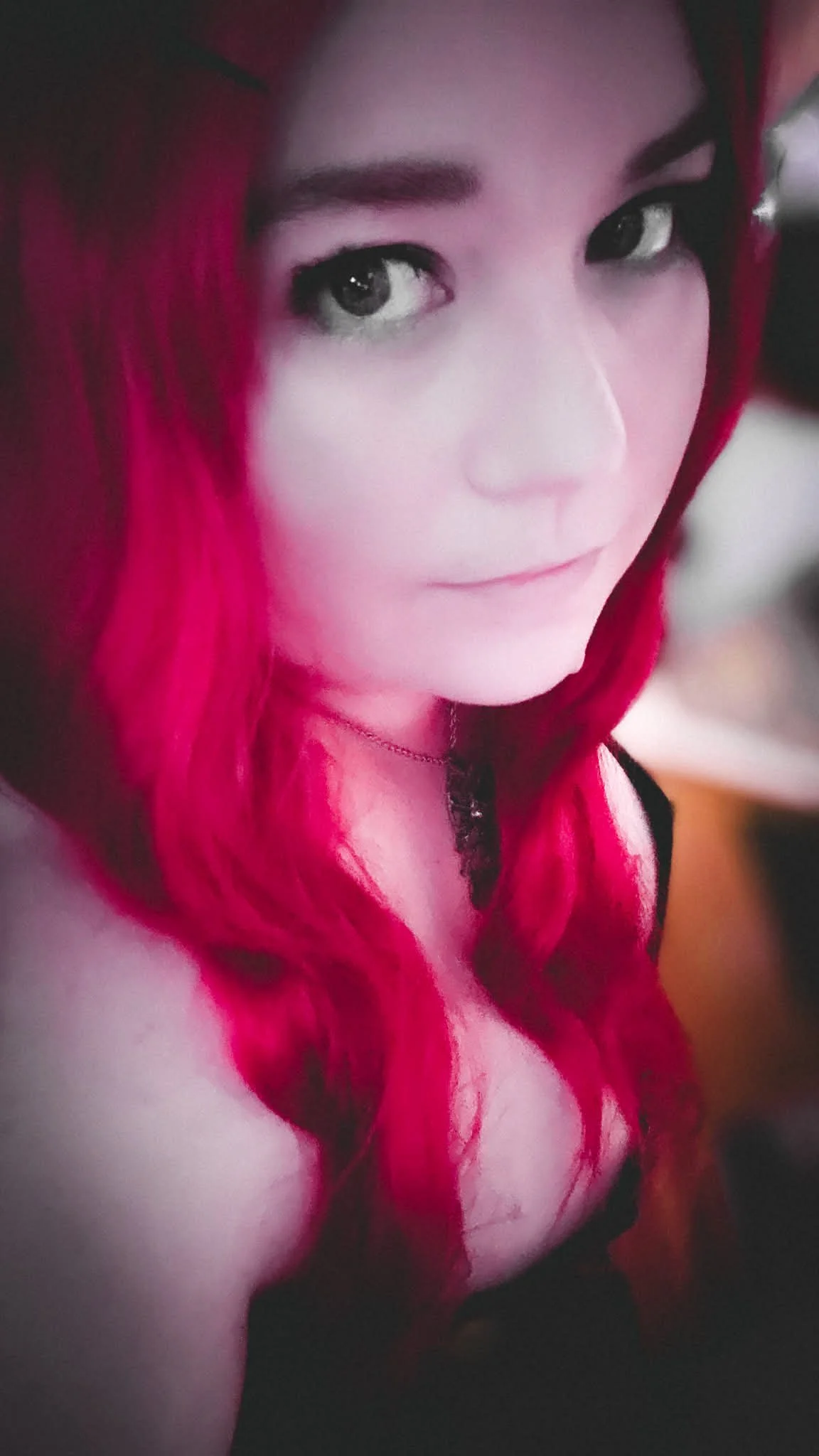 A young woman with bright red hair styled in loose waves. She has striking dark eyes and is wearing light makeup. She is looking at the camera with a slight smile, wearing a black top and a delicate necklace.