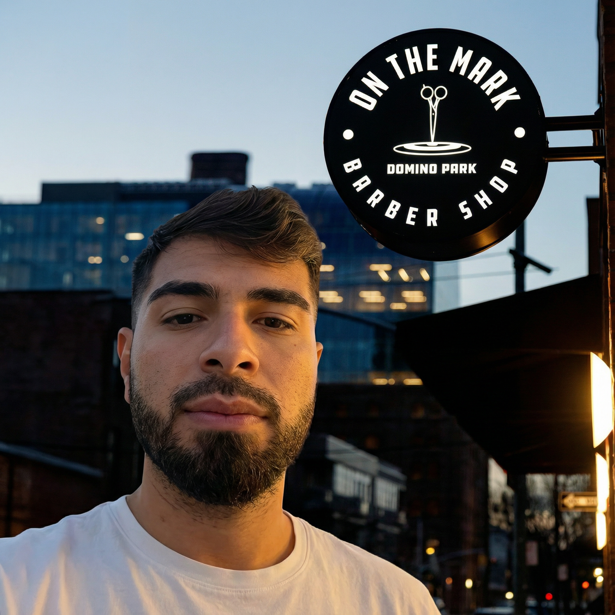 Jack, barber at On The Mark Barbershop in Domino Park, Brooklyn, standing outside the shop with the illuminated On The Mark sign.