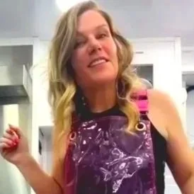 Woman with blonde hair wearing a purple apron, smiling in a kitchen.