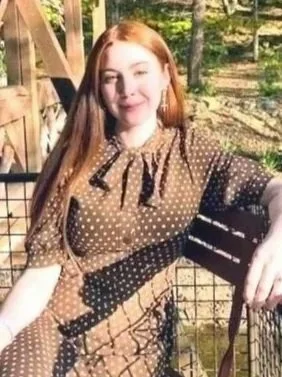 Young woman with long red hair outdoors near a wooden fence and trees, smiling at the camera.