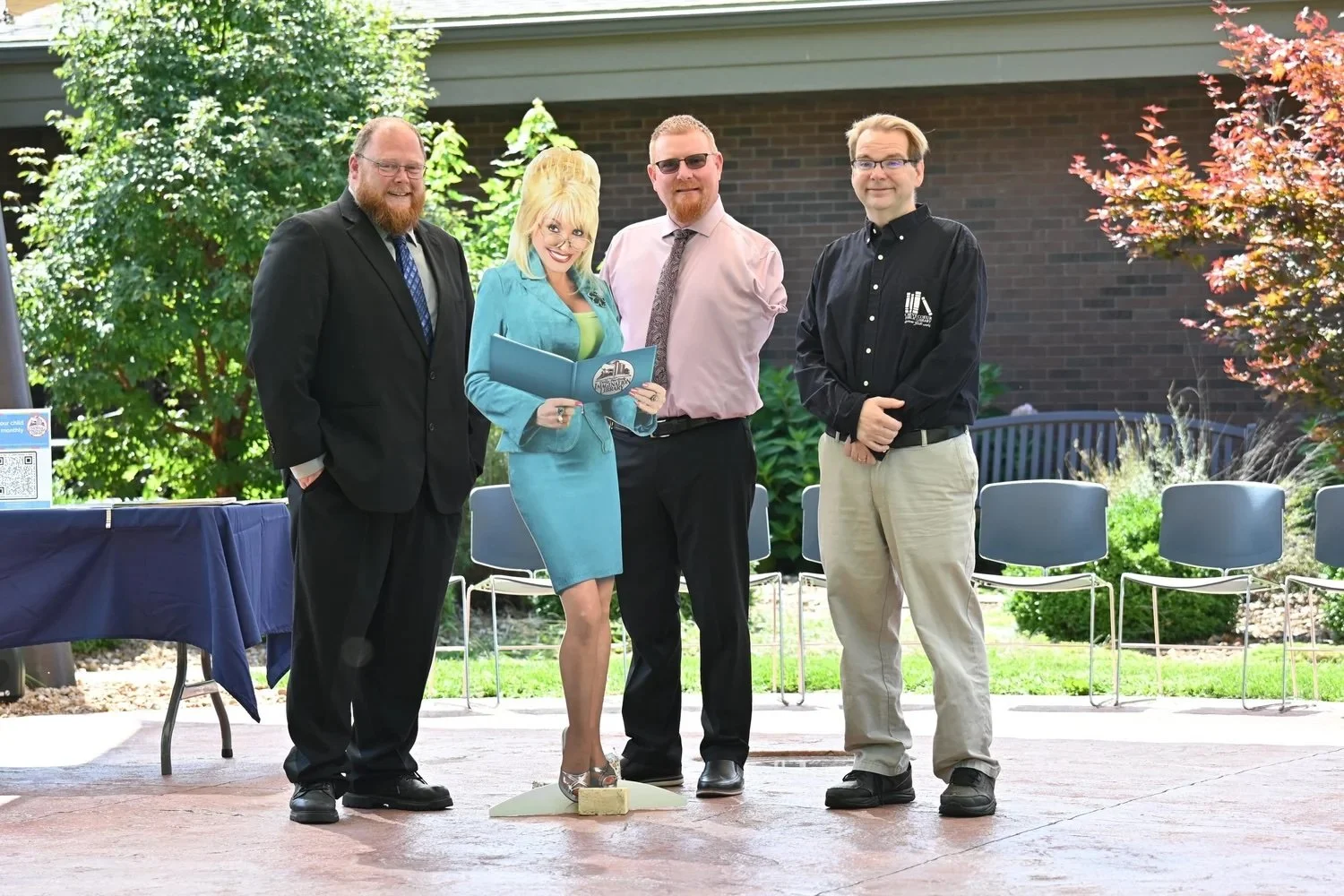 Three men formally dressed, smiling next to a cut-out of Dolly Parton reading a book