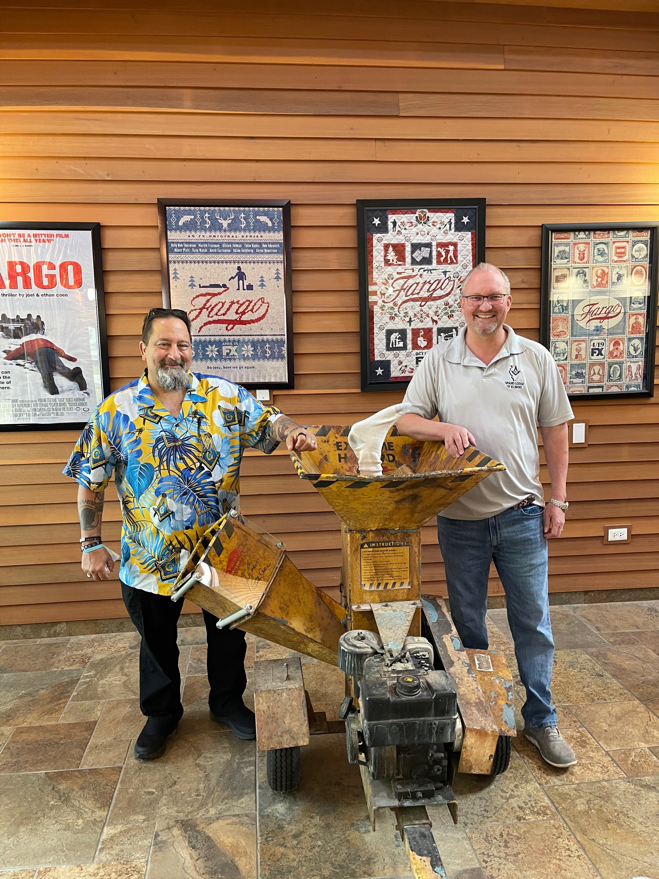 Two men pose and stand next to a prop from the movie Fargo, with multiple Fargo posters framed on the wall behind them
