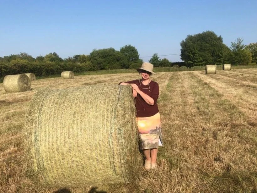 A woman wearing a hat and brown shirt leaning on a large rolled hay bale in an open field with more hay bales and trees in the background on a sunny day.