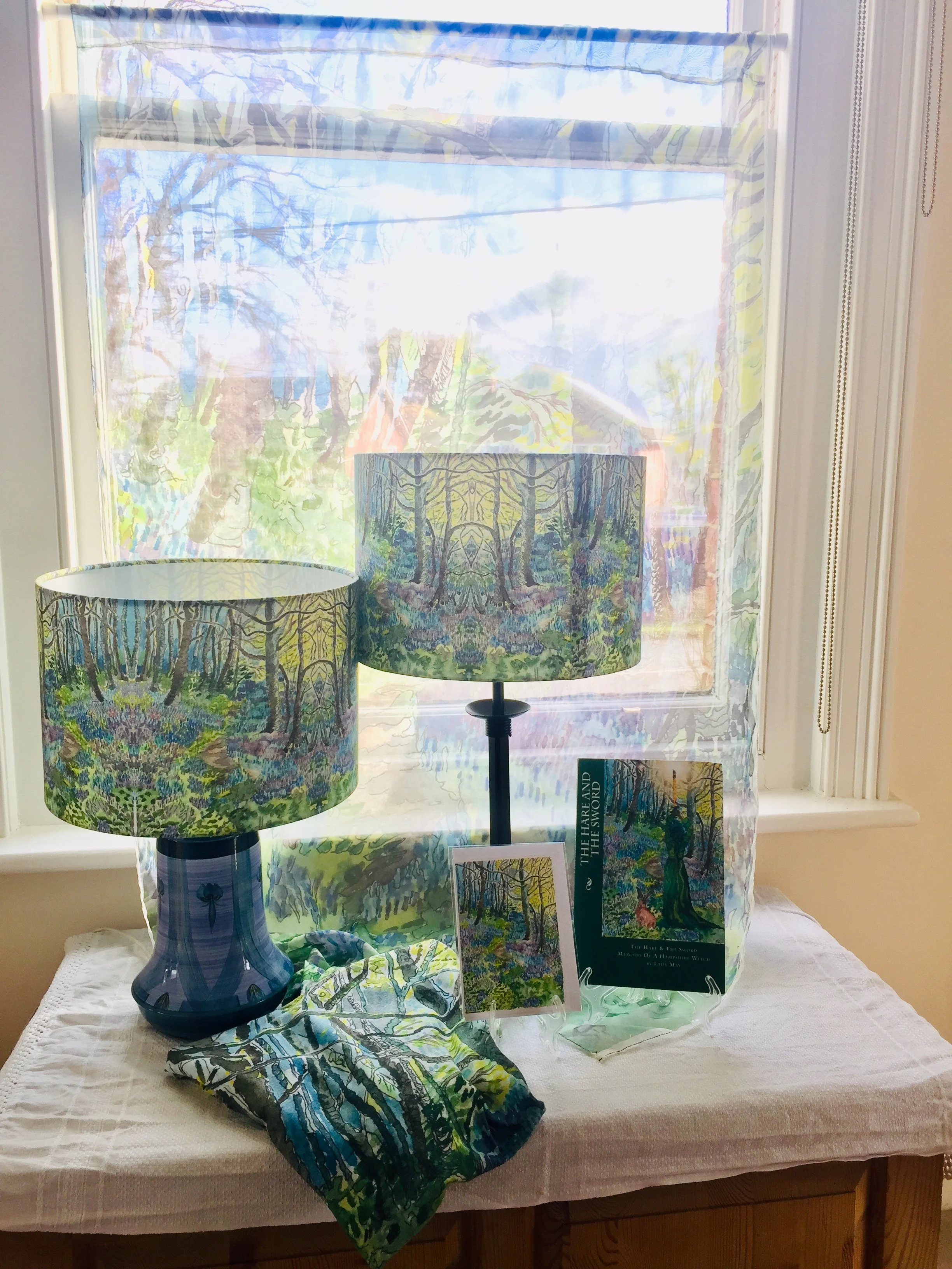 Decorative lamps with forest-themed shades, a book titled 'The Hare and the Sword,' and artwork featuring a forest scene on a table near a window with sheer curtain.