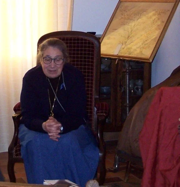 An elderly woman with glasses, wearing a black sweater and blue skirt, sits on a wooden chair with a high backrest, hands clasped. There is a map or antique document on an inclined stand behind her and part of a bed with a red blanket on the right.