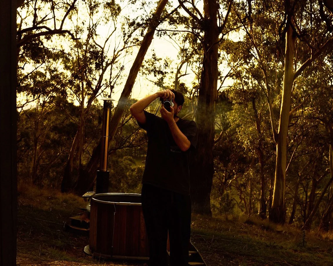 A person taking a photo in a wooded outdoor area during sunset or sunrise with large trees and a hot tub in the background.