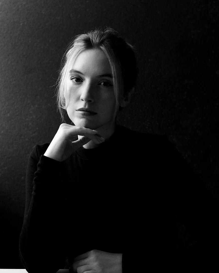 A black and white portrait of a woman with light hair, resting her chin on her hand, looking directly at the camera against a dark textured background.