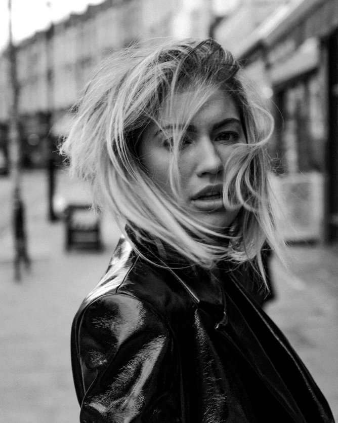 Black and white photo of a woman with tousled blonde hair wearing a shiny black leather jacket, standing outdoors in an urban environment.