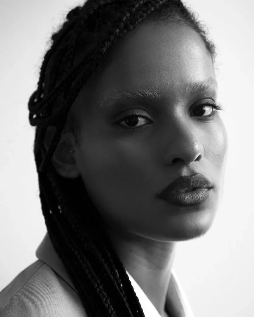 Close-up black and white portrait of a woman with braided hair, wearing a collared shirt, looking at the camera with a neutral expression.