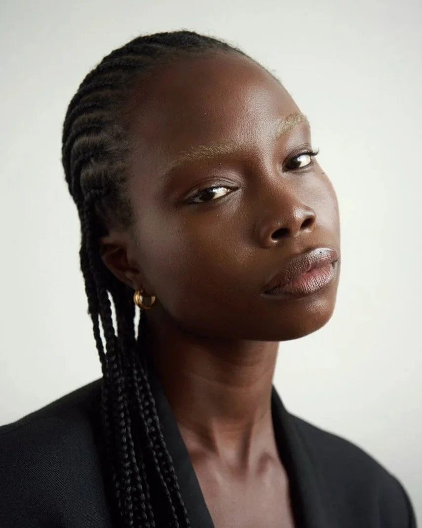 Close-up portrait of an African American woman with braided hair, wearing a black blazer and gold hoop earrings, looking confidently at the camera against a plain light background.