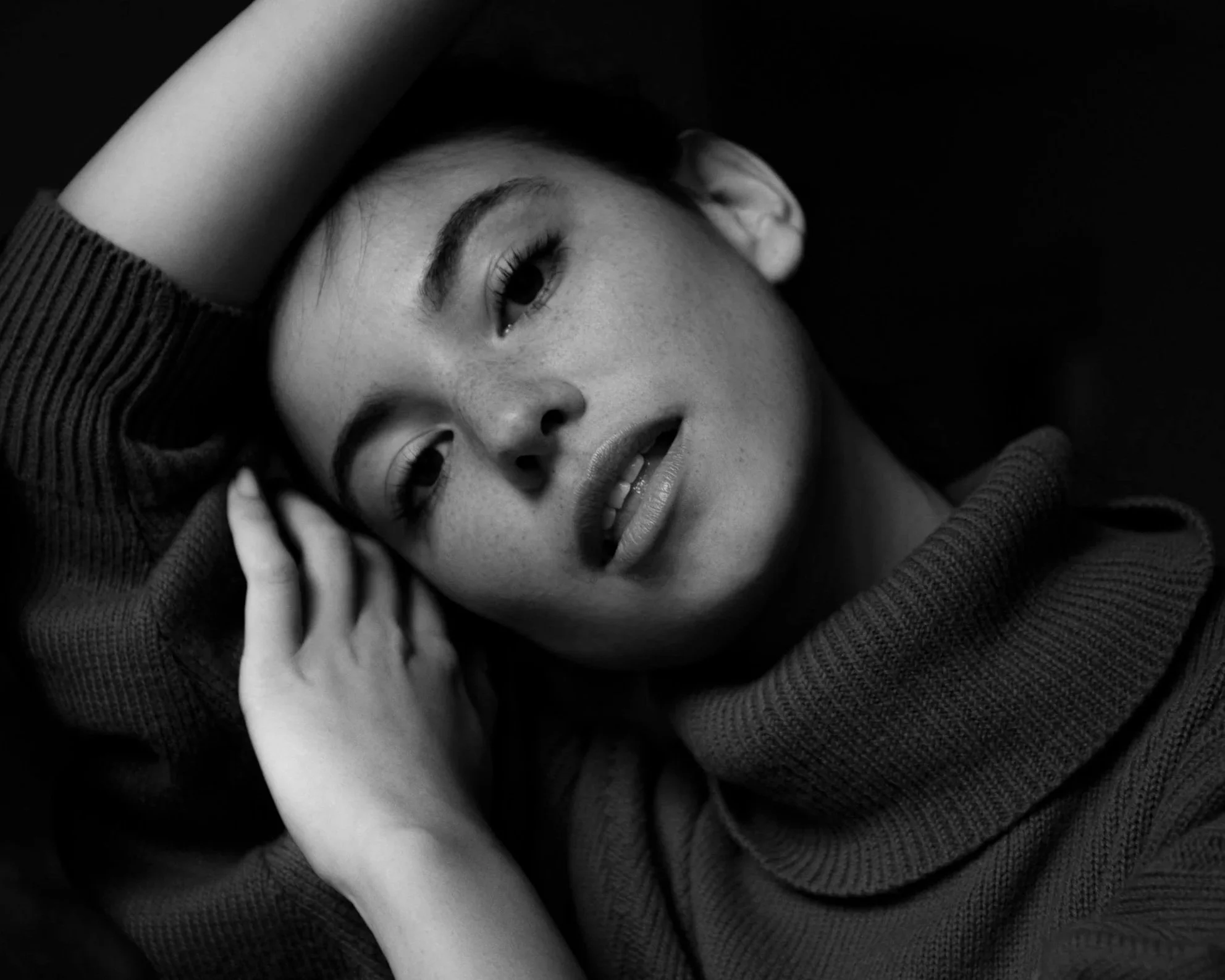 A black-and-white portrait of a woman lying down with her head resting on her arm, wearing a knitted sweater, with a calm expression, slightly open lips, and her eyes gazing to the side.