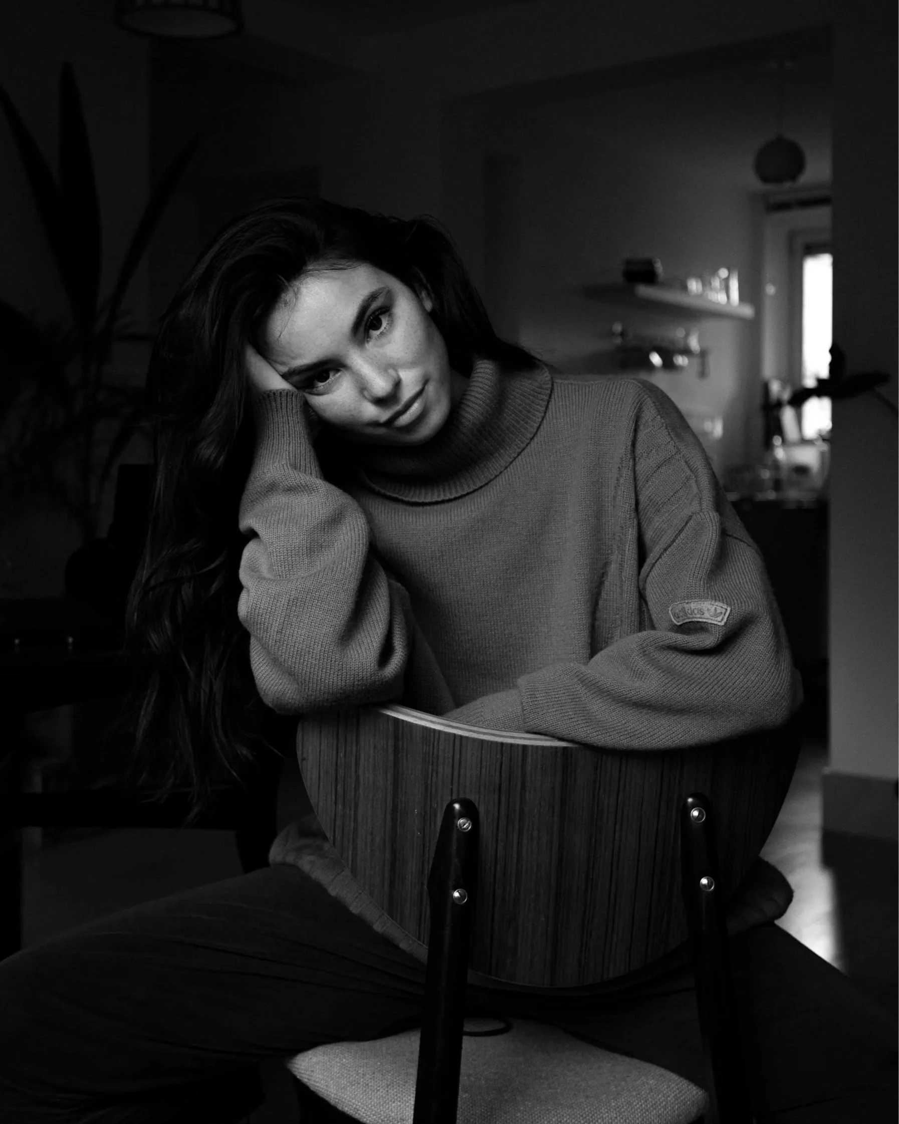 A black and white photo of a woman with long hair sitting at a table with a guitar, resting her head on her hand and looking directly at the camera in a cozy indoor setting.