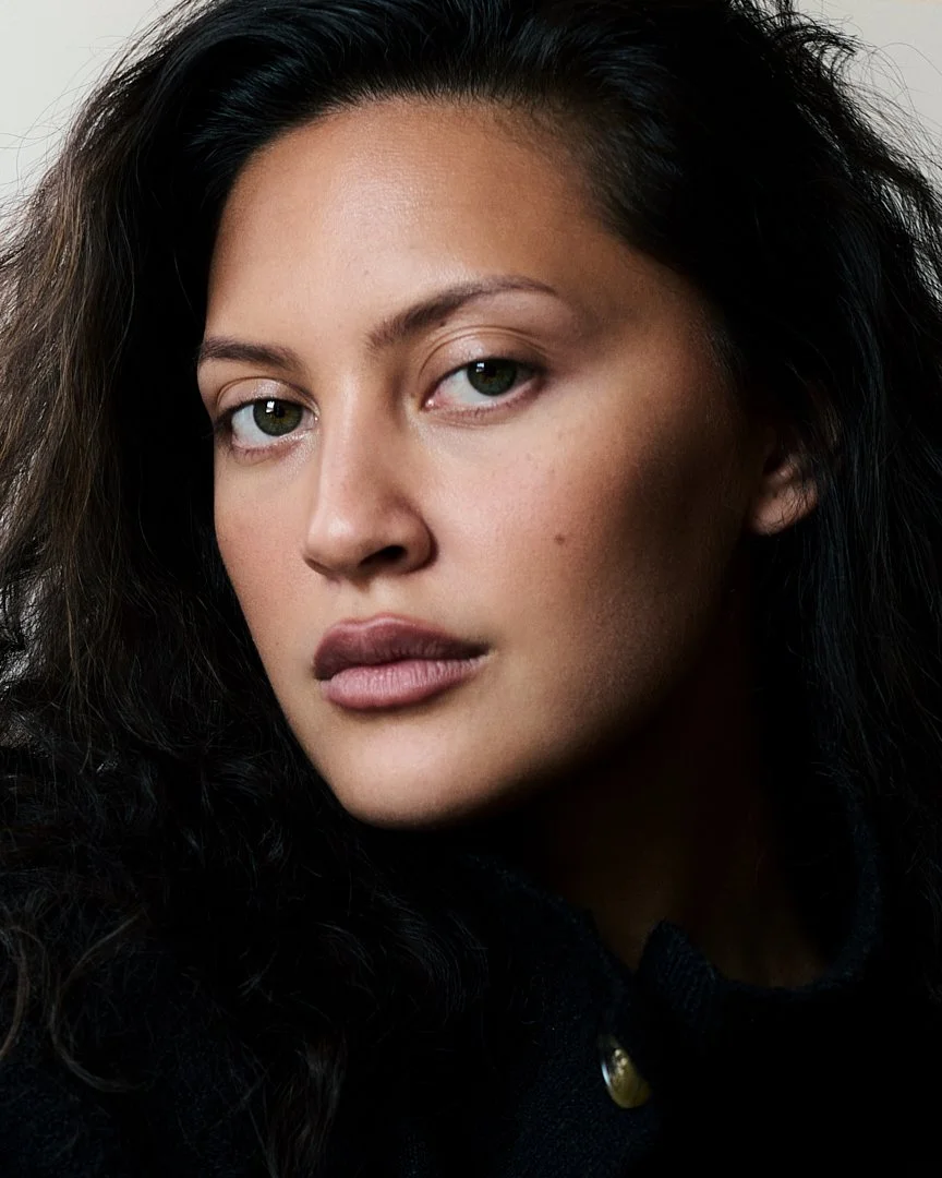 Close-up portrait of a woman with wavy dark hair and light green eyes, wearing a black top with a button, looking directly at the camera.