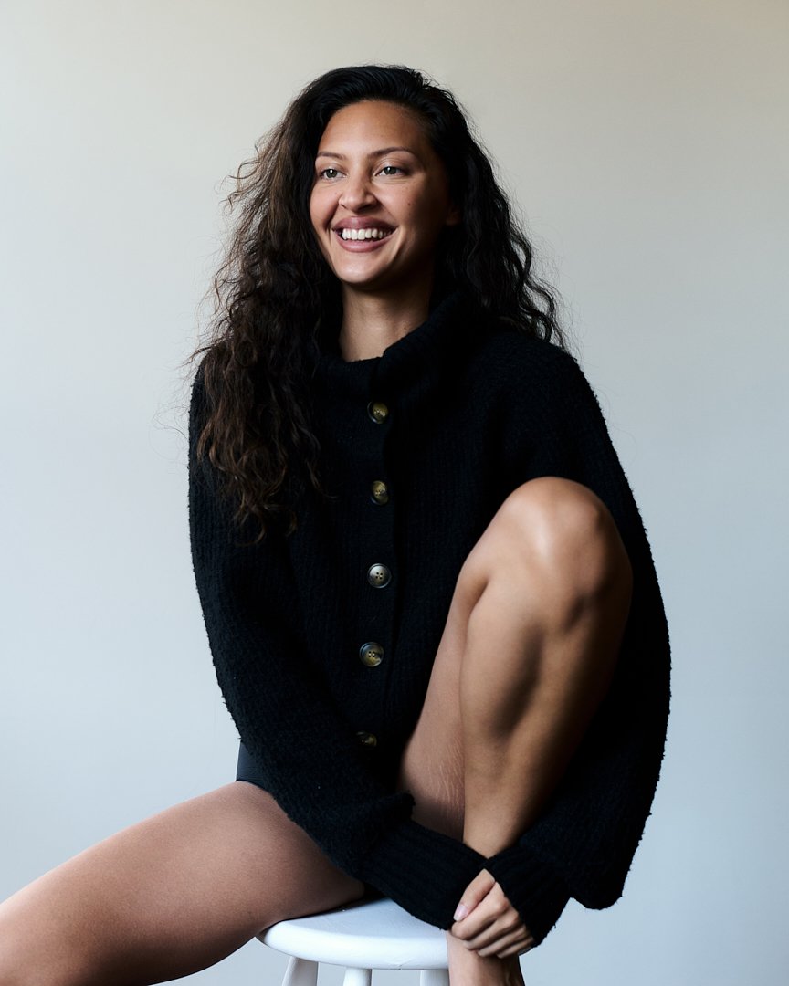 A woman with long curly dark hair, sitting on a white stool, smiling and wearing a black button-up sweater.