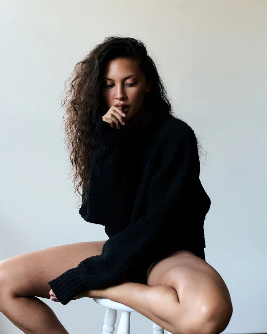 Woman with long, curly brown hair wearing a black sweater, sitting on a white stool against a plain light-colored background.