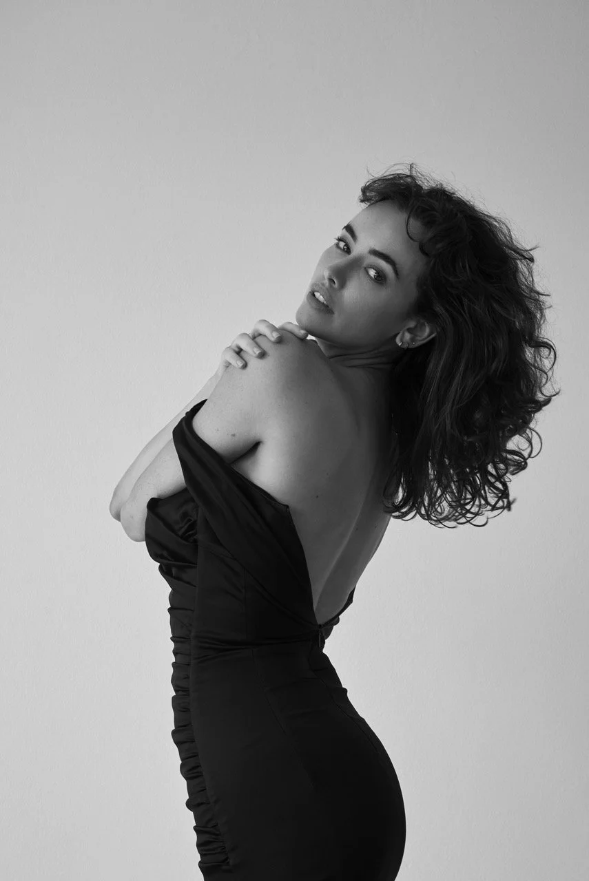 Black and white portrait of a woman with curly hair, posing with her arms crossed over her chest, wearing a strapless dress, against a plain background.