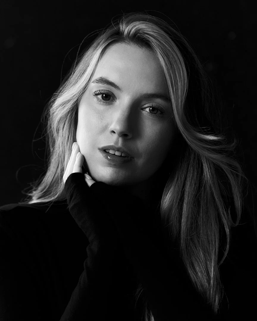 Black and white portrait of a young woman with long wavy hair, looking directly at the camera, touching her face with one hand, wearing dark clothing.