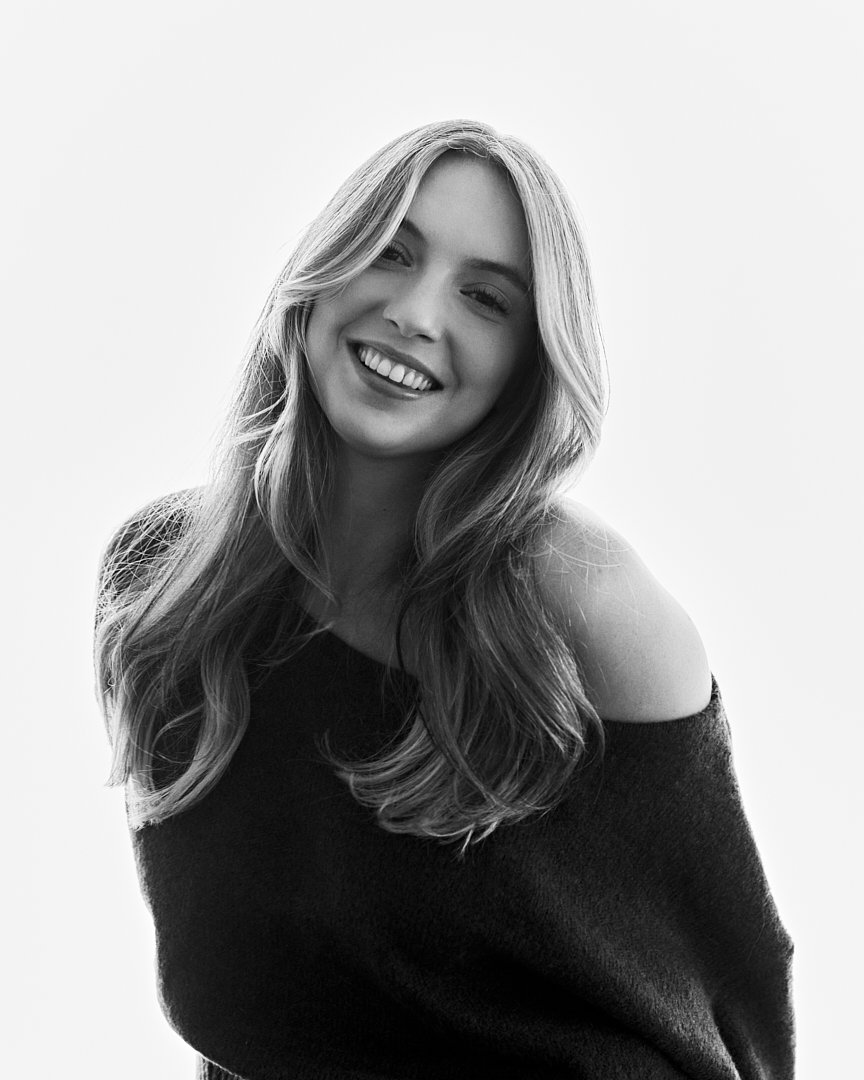 Black and white portrait of a smiling woman with long, wavy hair wearing a off-shoulder sweater against a plain background.