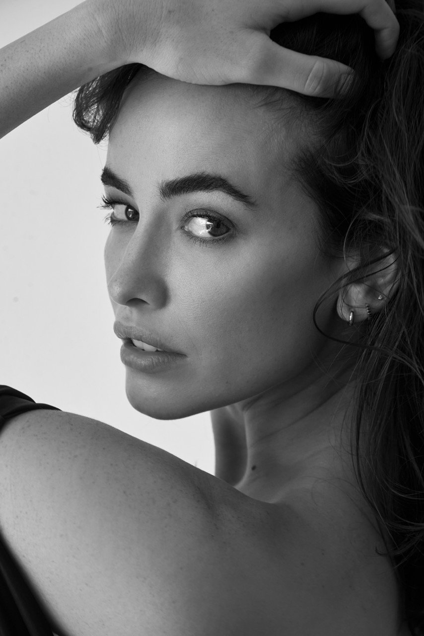 Black and white close-up portrait of a woman with her hand in her hair, showing her face, eye, and shoulder, wearing earrings.