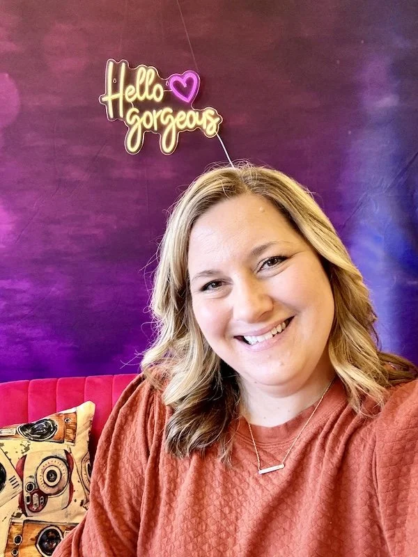 Nicole taking a selfie in front of a purple wall with a neon sign that says 'Hello Gorgeous' with a heart. She is smiling, has shoulder-length wavy hair, and is wearing a red top and a simple necklace.