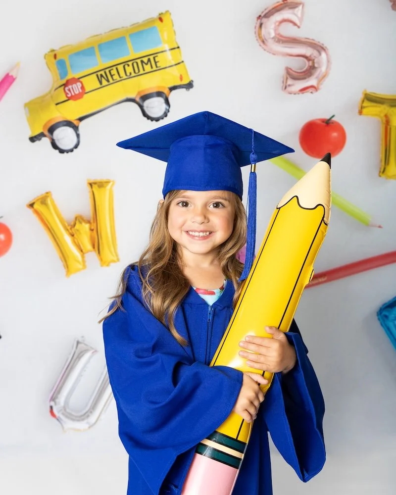 Girl in blue graduation cap and gown holding large yellow pencil shaped inflatable, smiling, with colorful balloon letters and images of a school bus, a red apple, and balloons in the background. Professional School Photographer