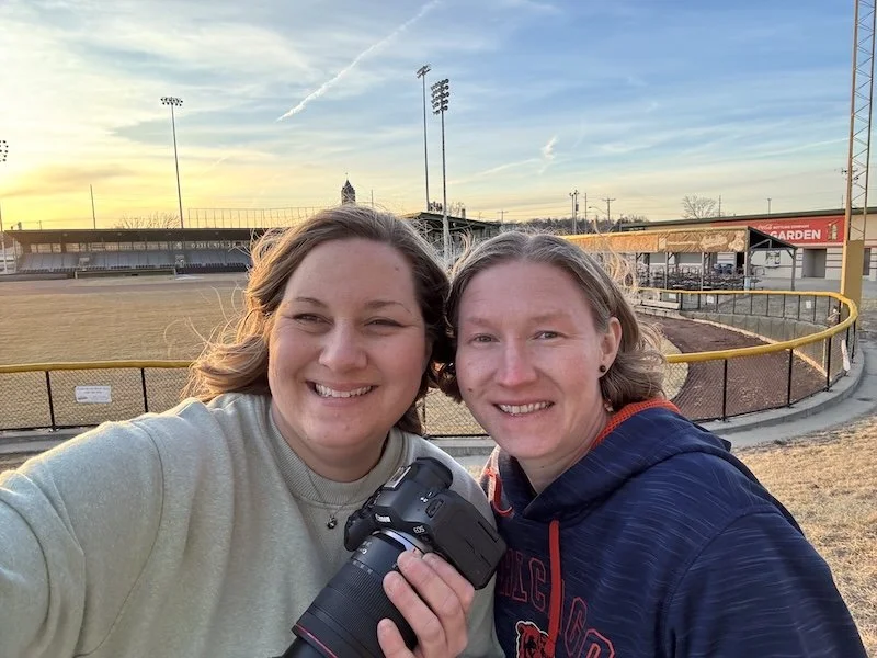 Two women smiling at the camera, standing on a sports field during sunset, with a camera in hand.