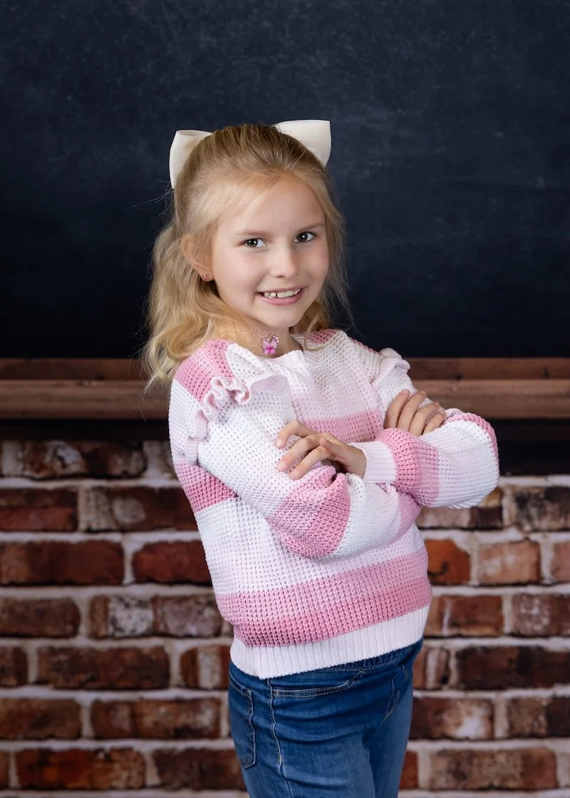 A young girl with blonde hair and a white bow, smiling with arms crossed, wearing a pink and white striped sweater and blue jeans, standing in front of a brick wall and chalkboard background.