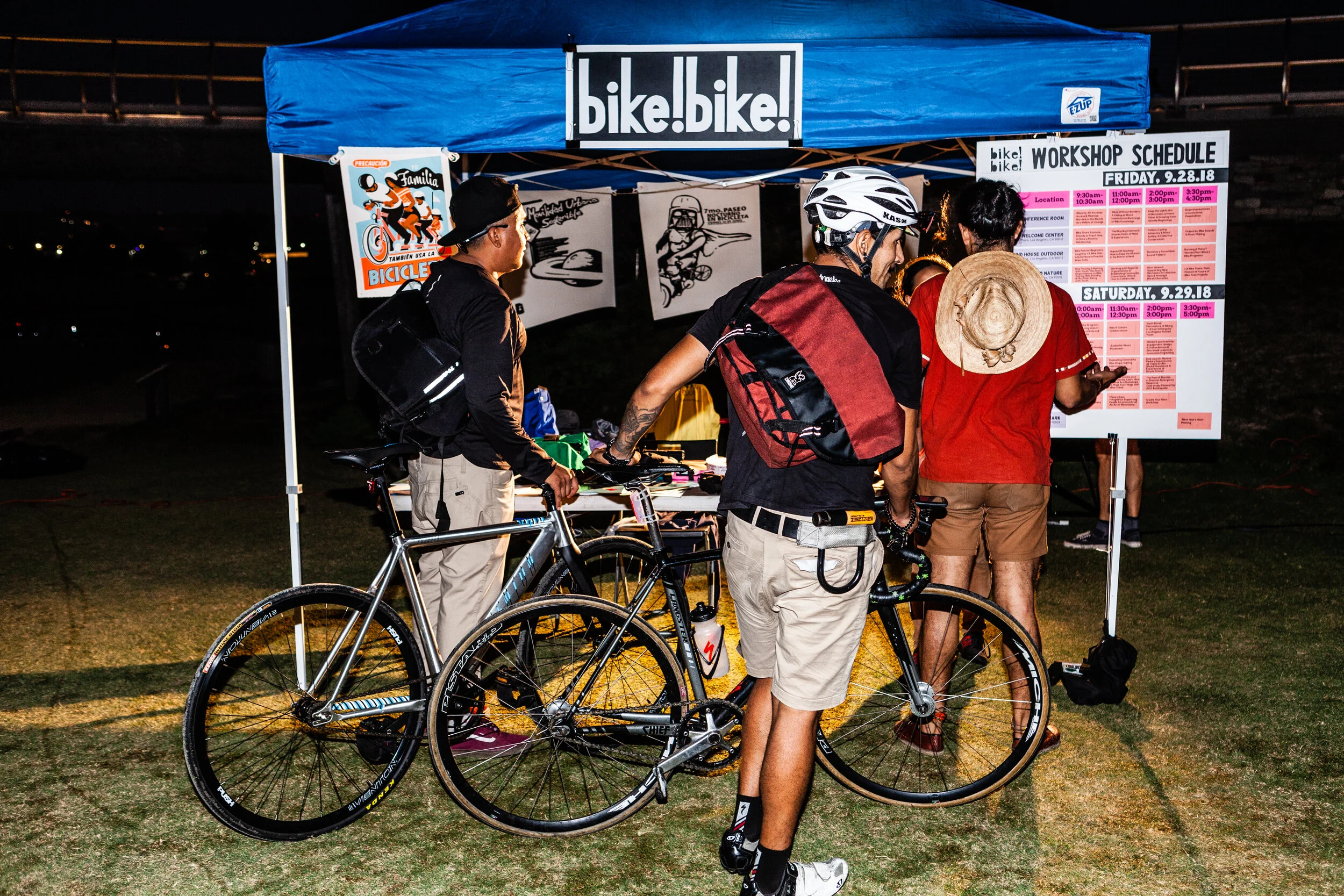 Three cyclists holding their bikes check out the Bike!Bike! 2018 Conference schedule posted on support beam of an outdoor pop-up tent. (Photo: Frank Hom - http://frankhom.com/)