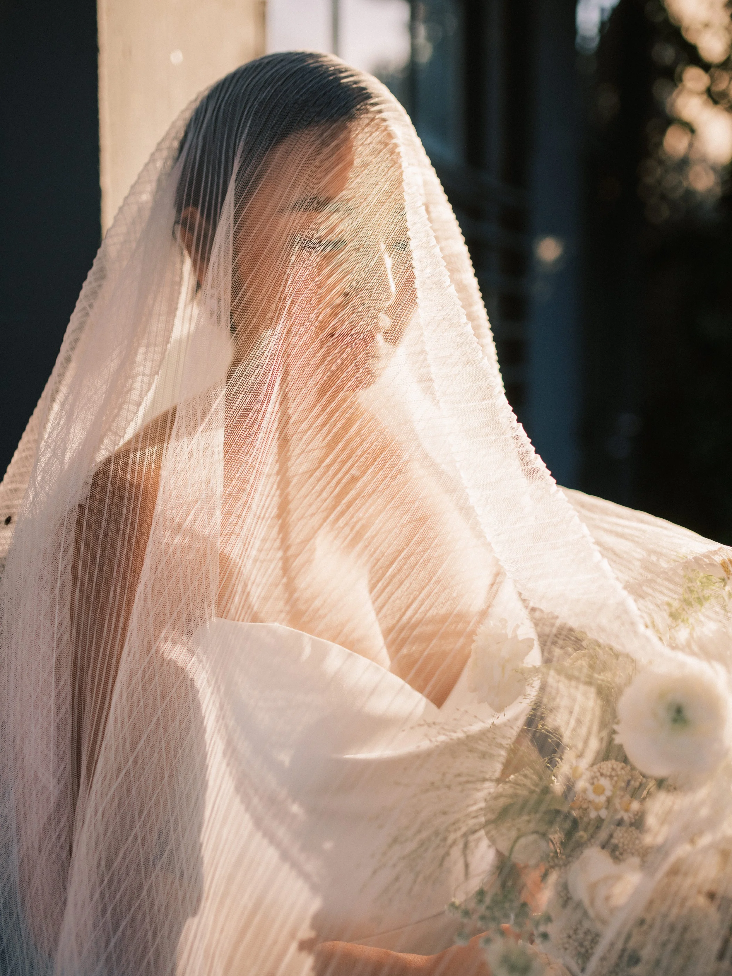 A woman with dark hair and glasses, covered with a sheer, pleated veil, standing outdoors near a building, with the sunlight illuminating her face and hair. | Luxury Wedding Planner, Southern California