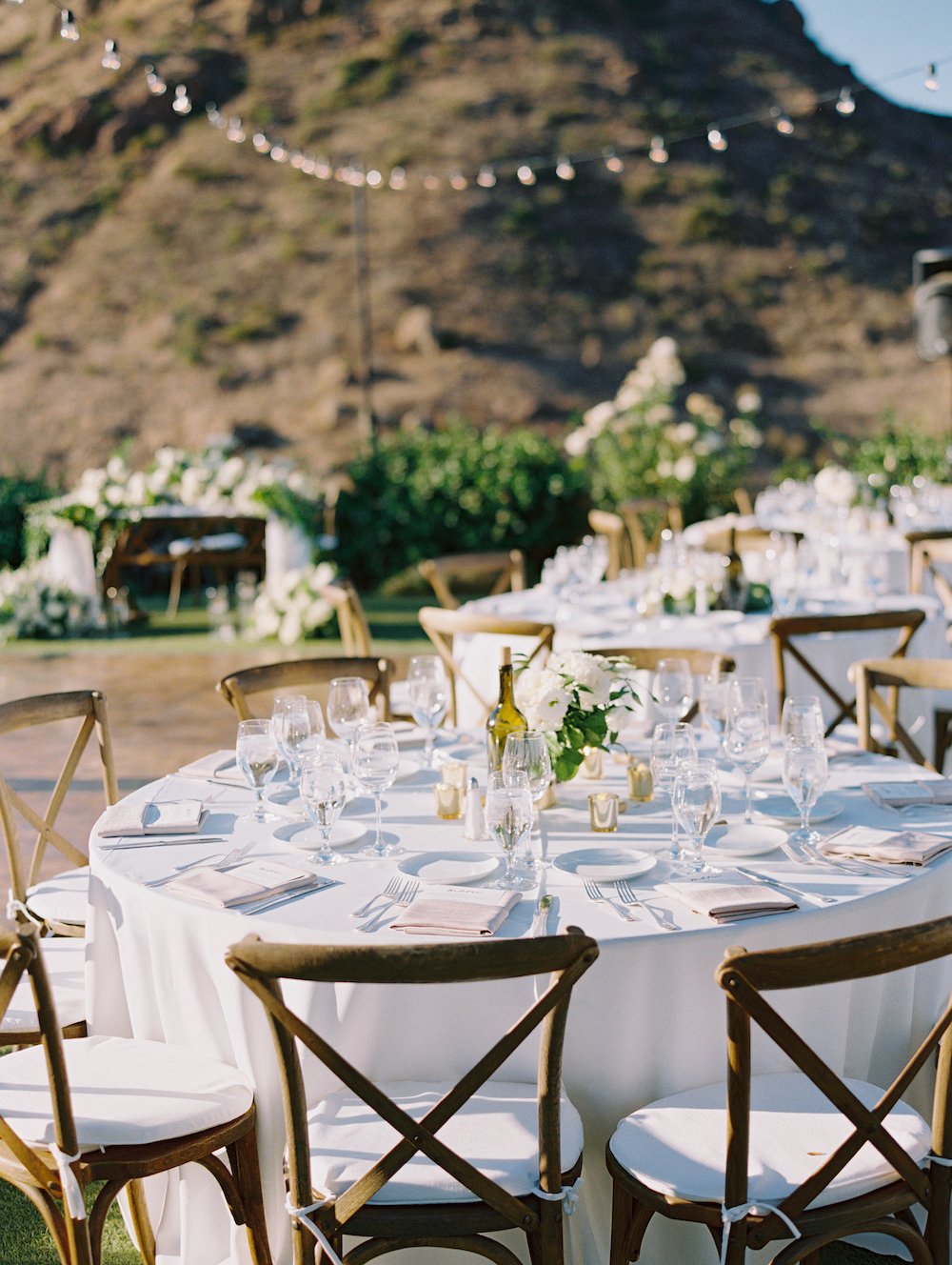 Outdoor wedding reception tables with white linens and wooden cross-back chairs.