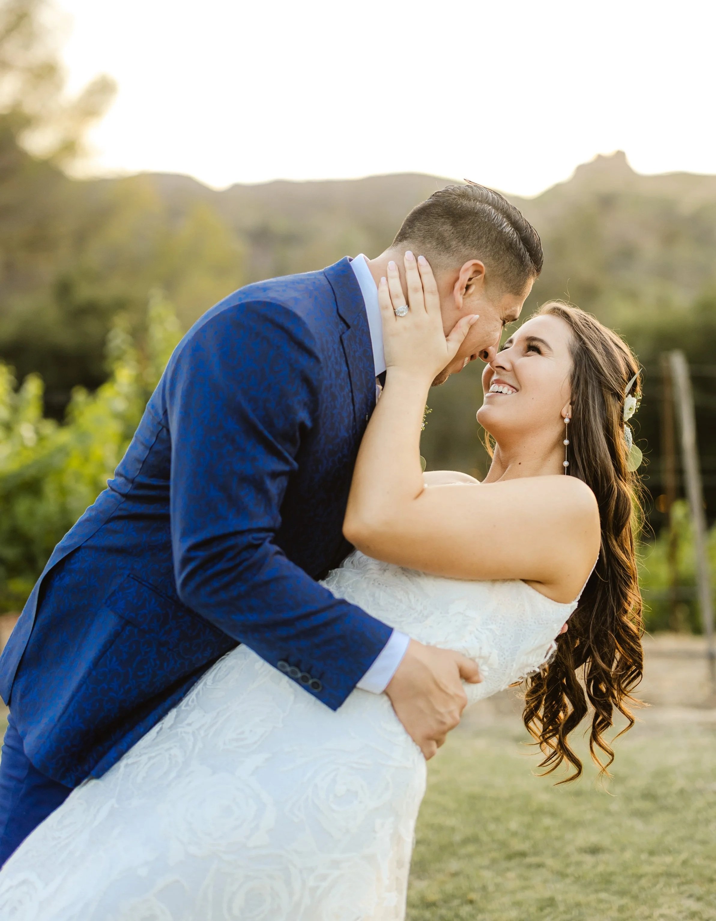 A bride and groom sharing a kiss outdoors, with the groom in a blue suit and the bride in a white lace dress, bright sunlight, and blurred greenery in the background. | Luxury Wedding Planner, Southern California