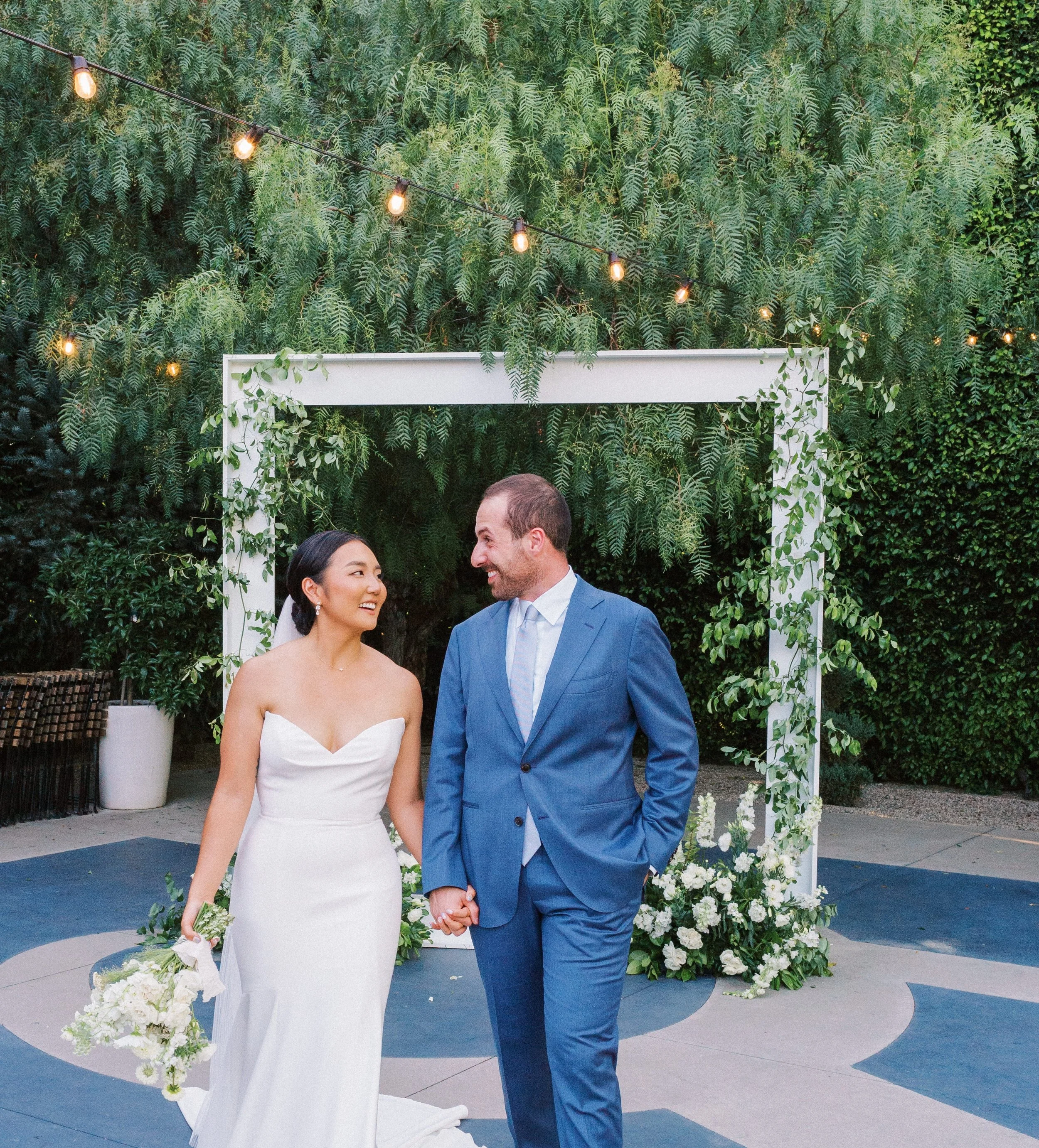 A bride and groom holding hands and smiling at each other outdoors during their wedding ceremony, with a white floral arch and string lights above them, surrounded by green foliage. | Luxury Wedding Planner, Southern California
