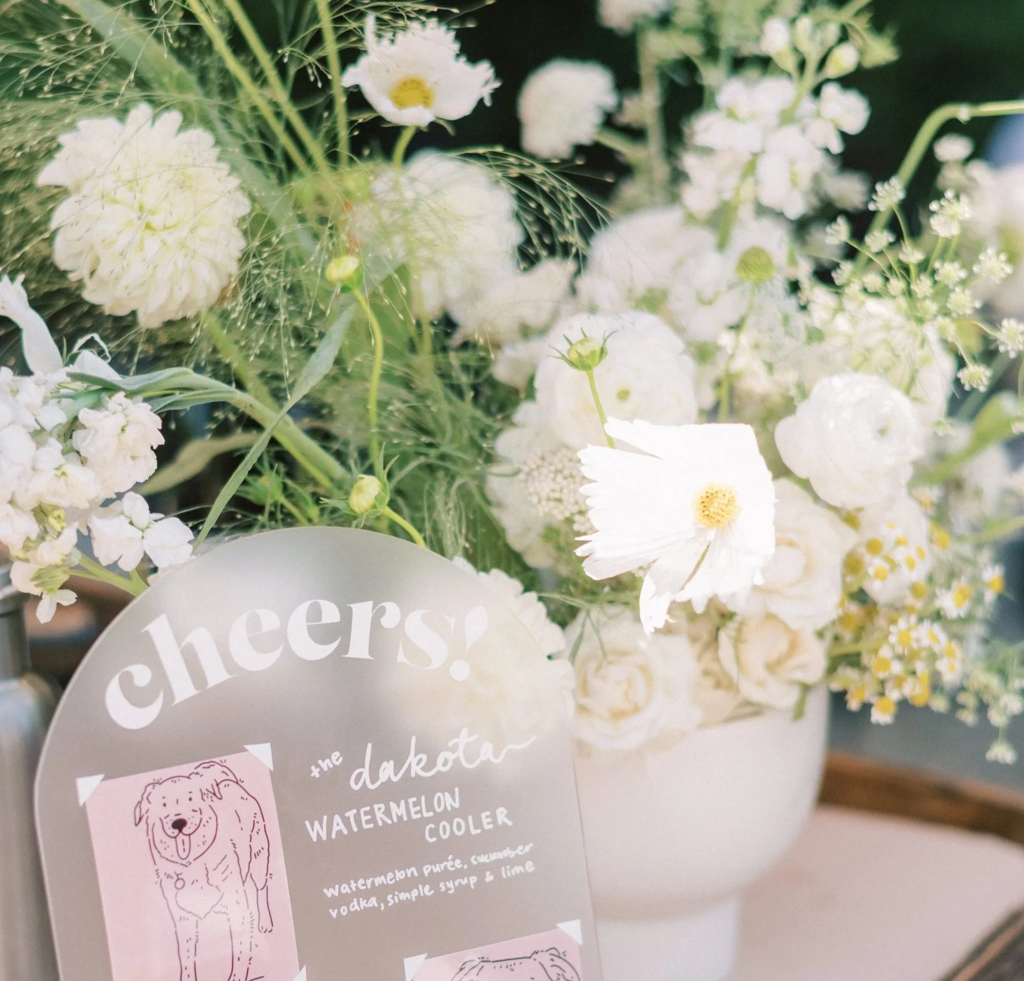 A floral arrangement with white flowers, green leaves, and a sign with a bear illustration listing ingredients for a drink including watermelon, cucumber, vodka, and lime. | Luxury Wedding Planner, Southern California