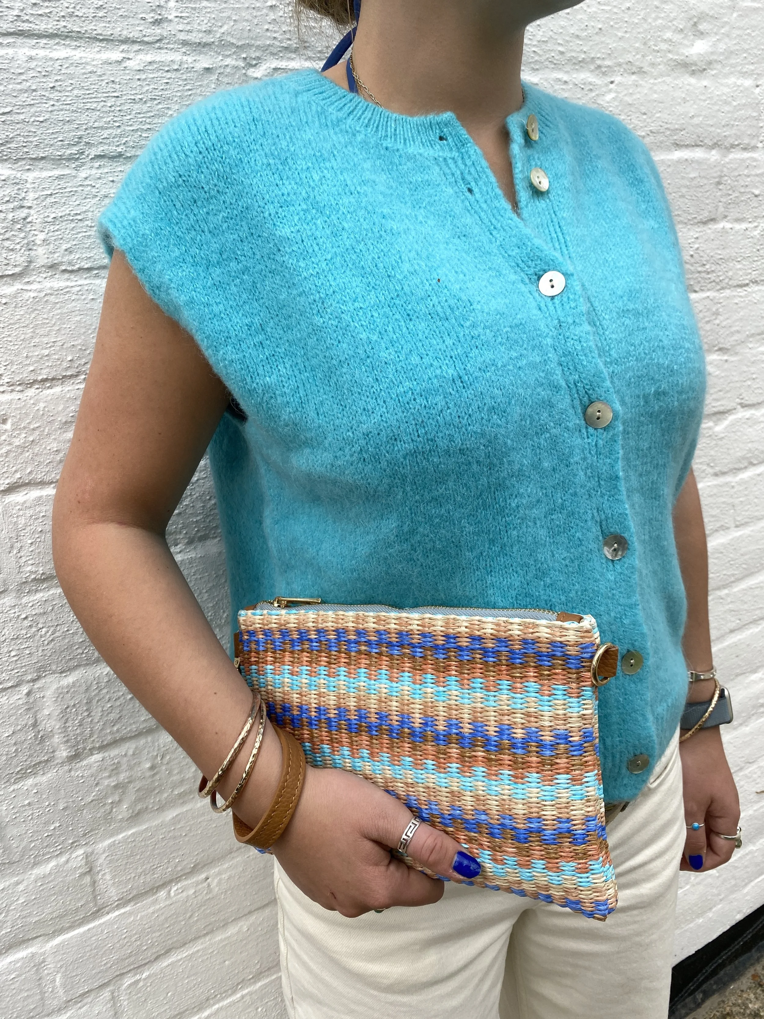 model wearing turquoise sleeveless cardigan and holding a raffia and leather clutch bag with blue and tan stripes