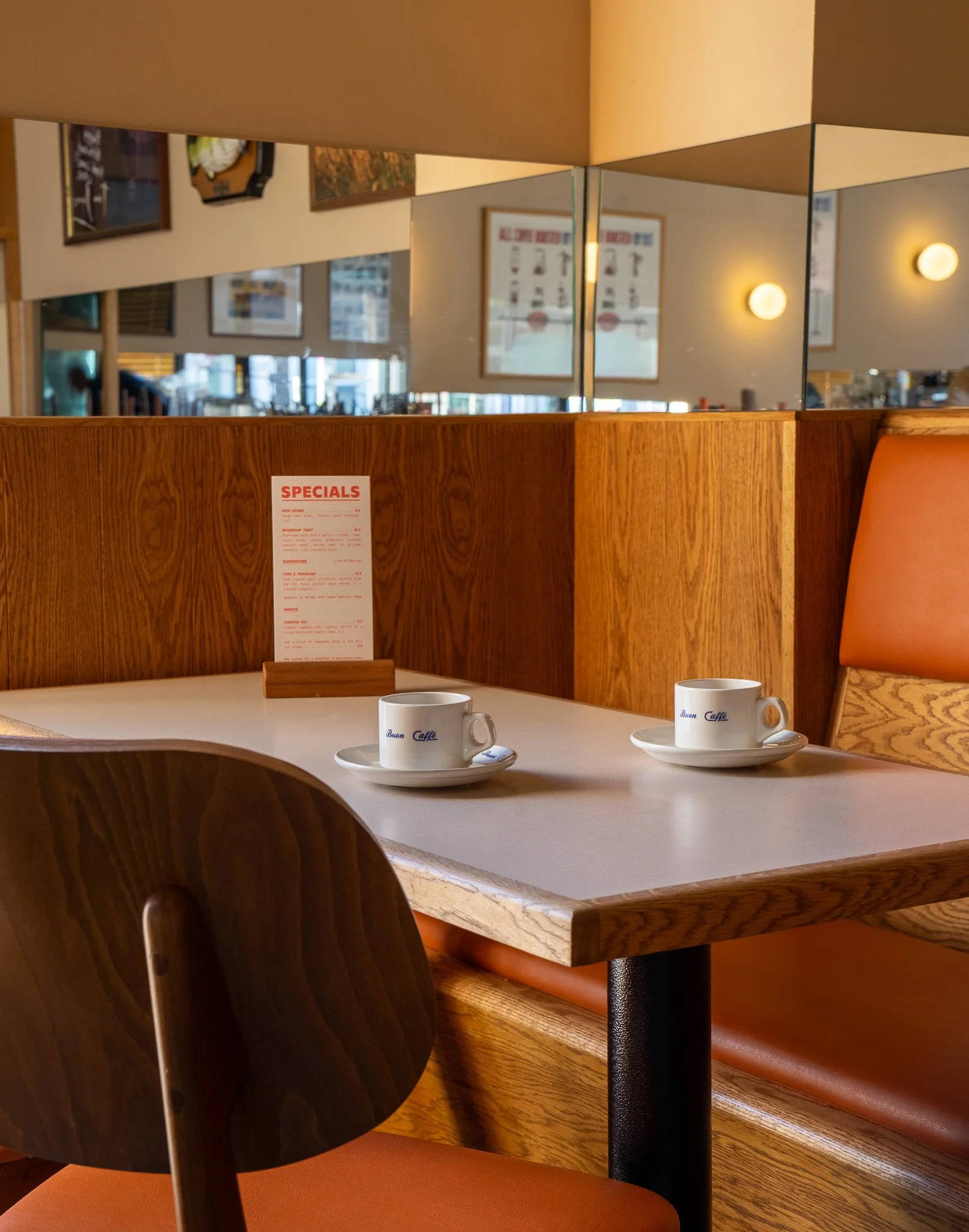 Two coffee cups on a table inside a cozy cafe, with a wooden booth and a framed menu in the background.