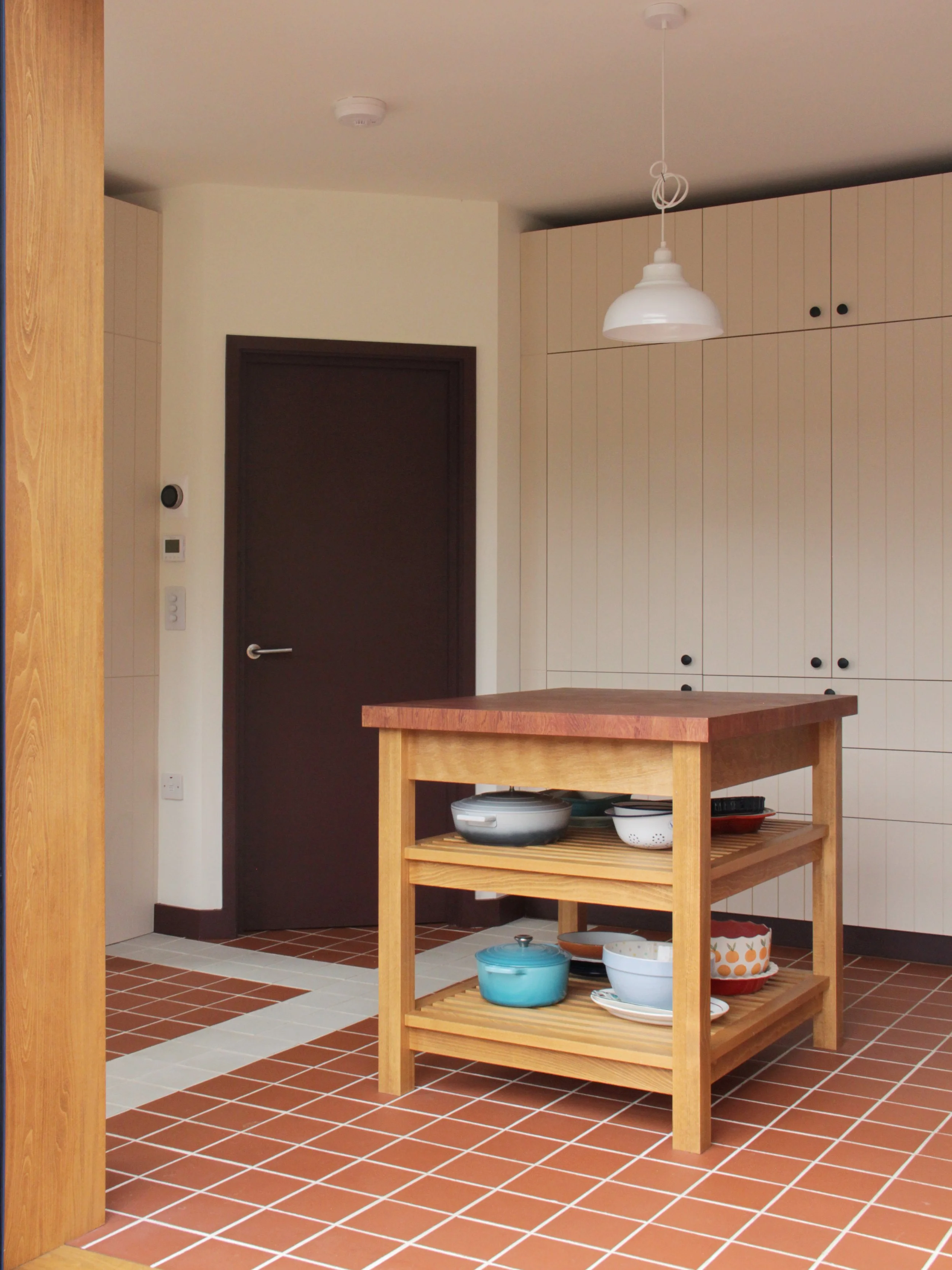 A kitchen with a small wooden island and a tiled floor, containing dishes and cookware on the shelves. There is a dark door, white cabinets, and a hanging pendant light.