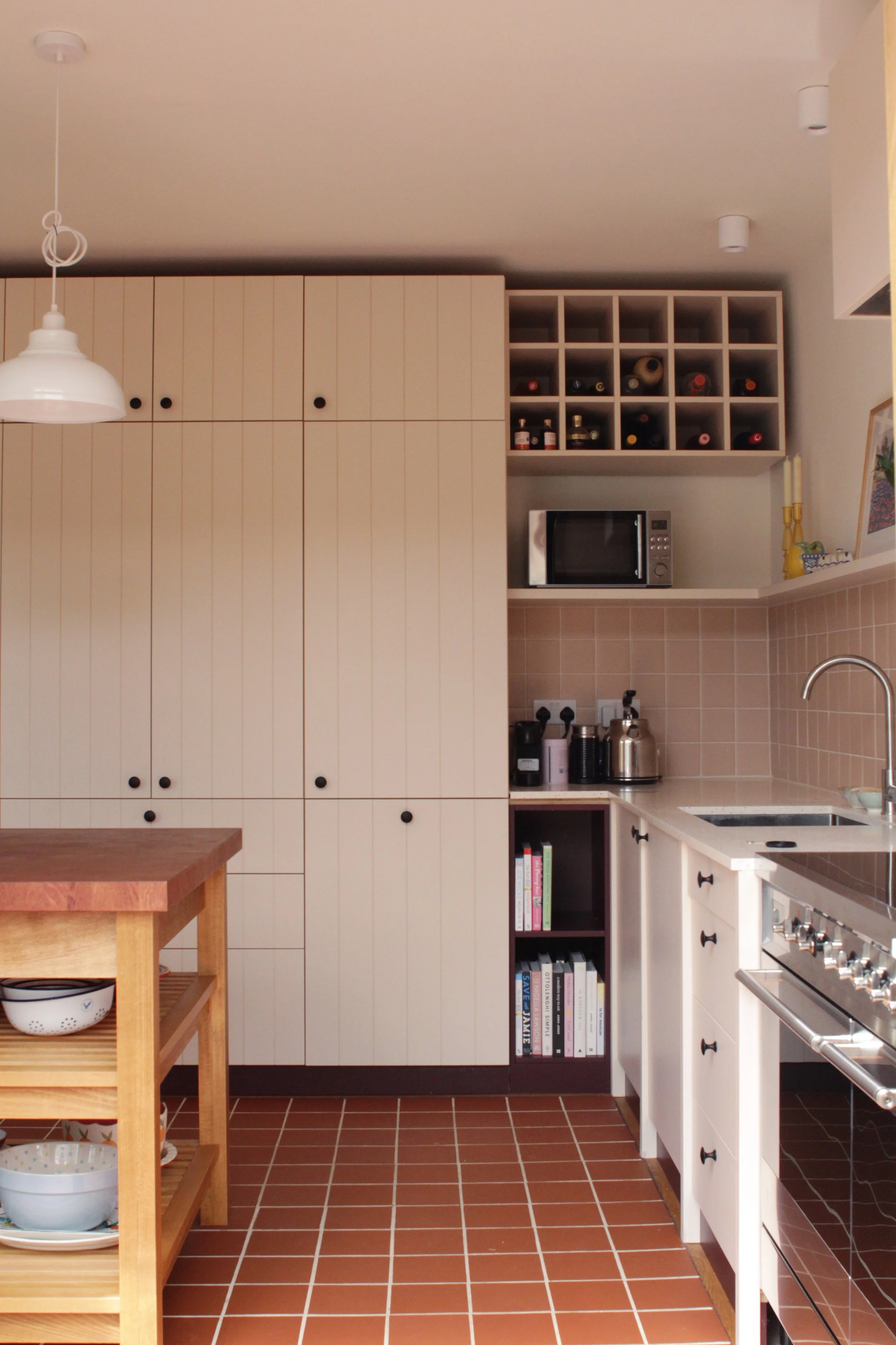 Kitchen with beige cabinets, a small open fridge, microwave, books, and kitchen utensils on beige tile countertops. Wooden kitchen island with bowls, terracotta floor tiles, and a white ceiling with hanging light.