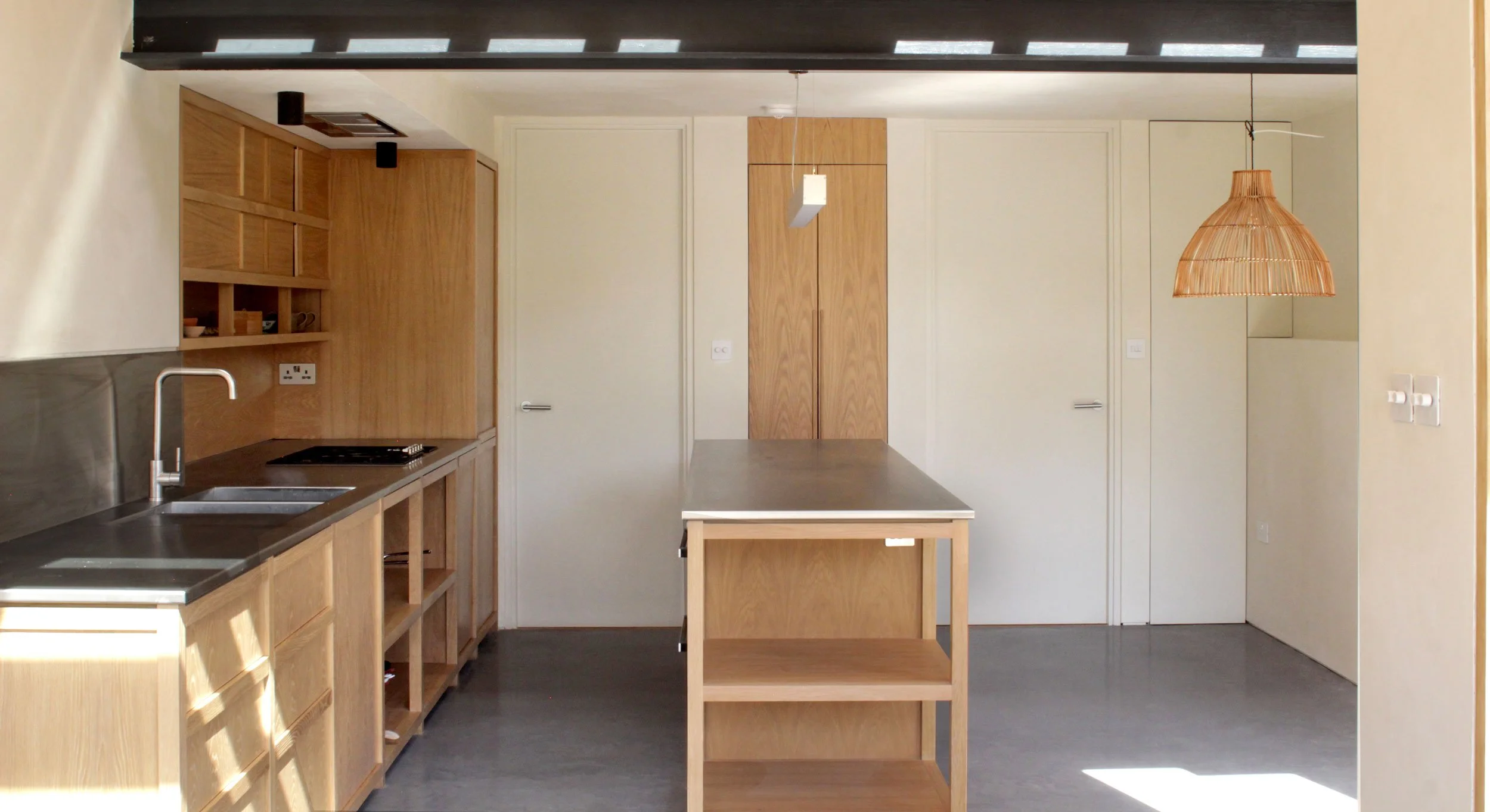 Modern kitchen with wooden cabinetry, open shelving, a black countertop, a stainless steel sink, a stove, a central wooden island with a gray countertop, a hanging wicker light fixture, and a white door.