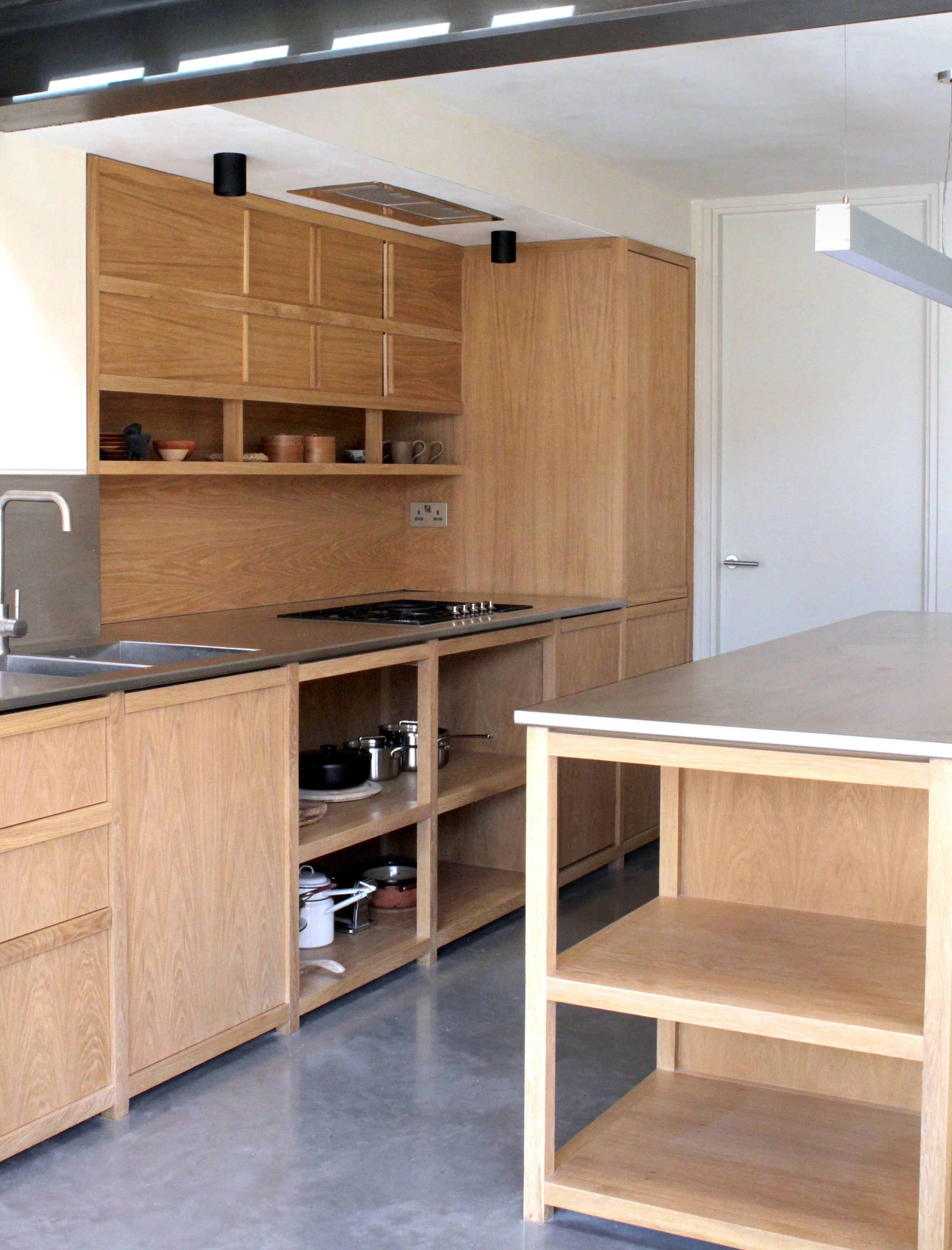 Kitchen with wooden cabinets, open shelves, and stainless steel sink, located in a modern home, with a white door and concrete floor.