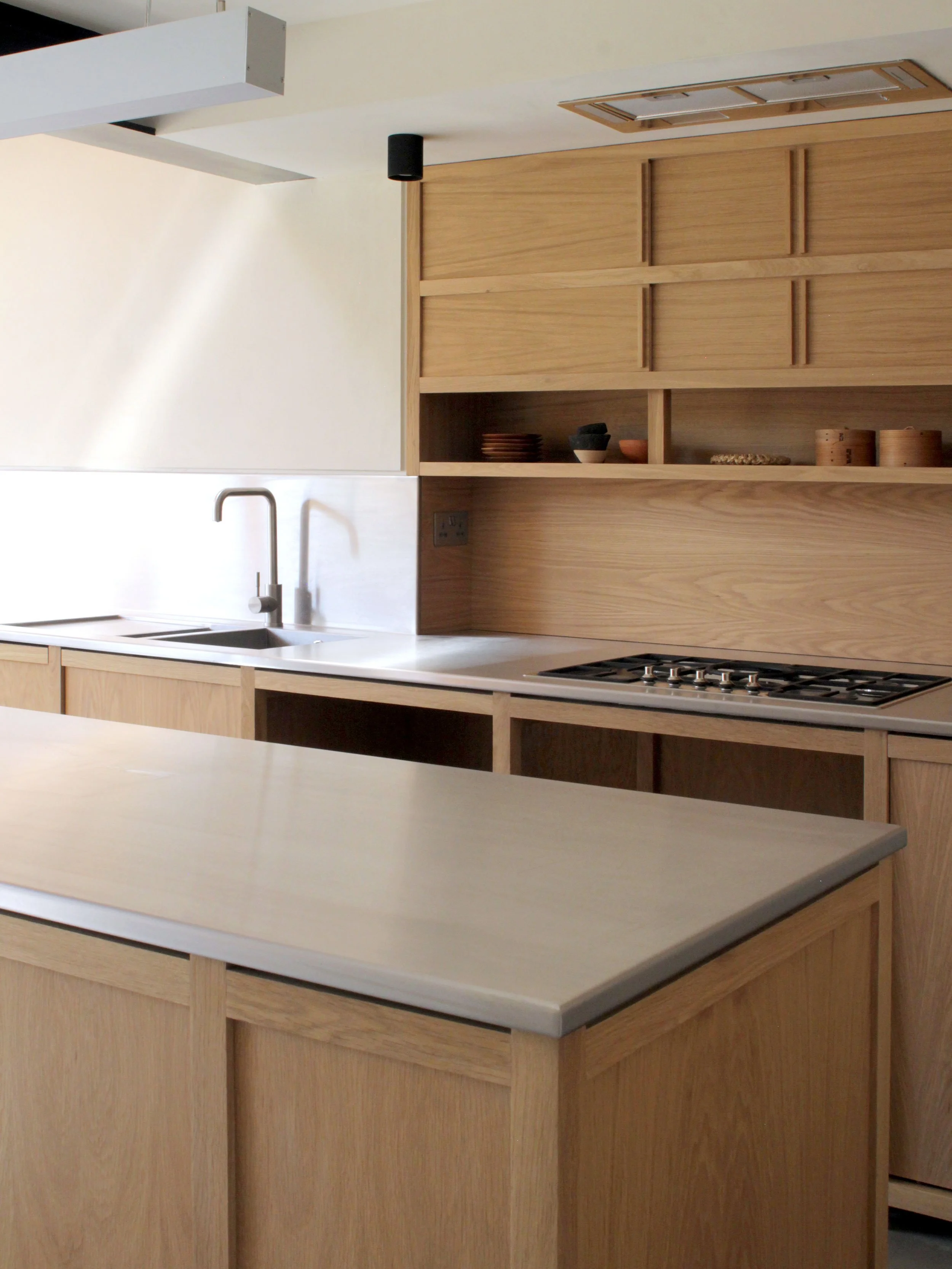 Modern kitchen with light wood cabinets, a white countertop, a stainless steel sink, and open shelves with pottery items.