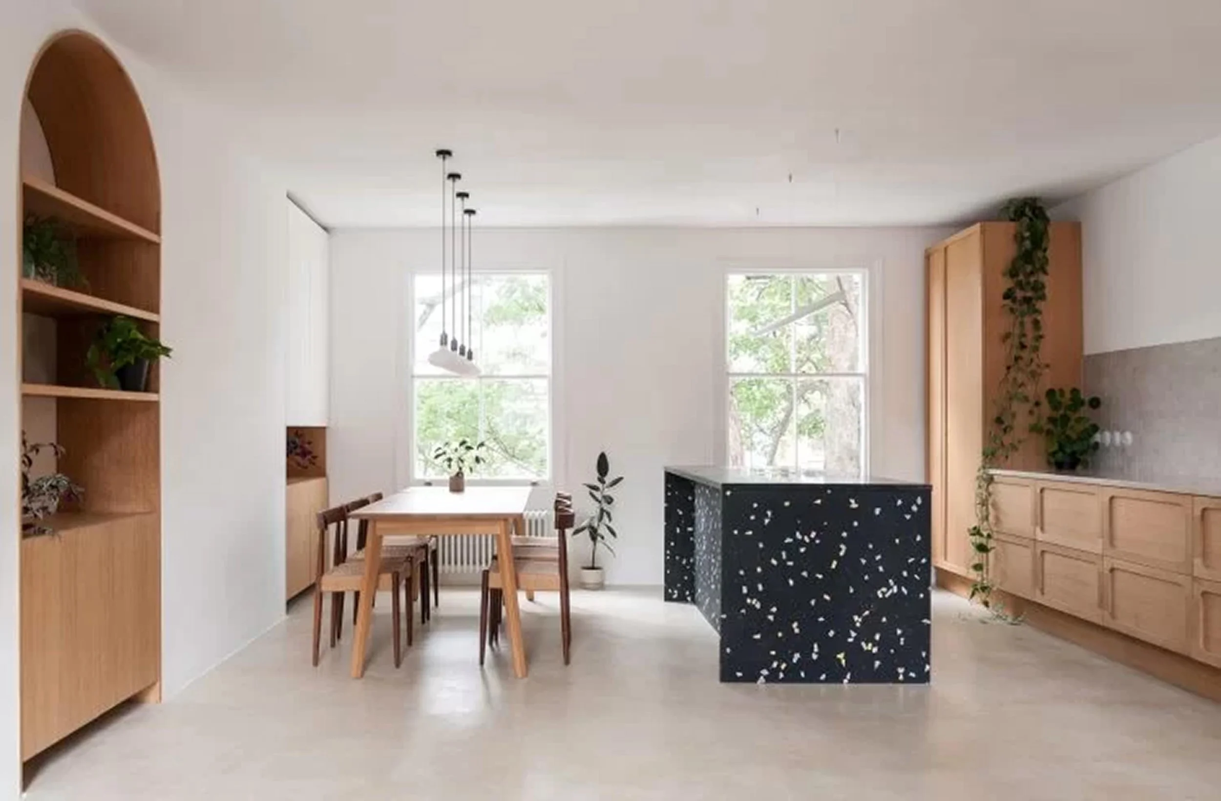 Modern minimalist kitchen and dining area with natural wood furniture, black terrazzo island, and green plants near large windows.