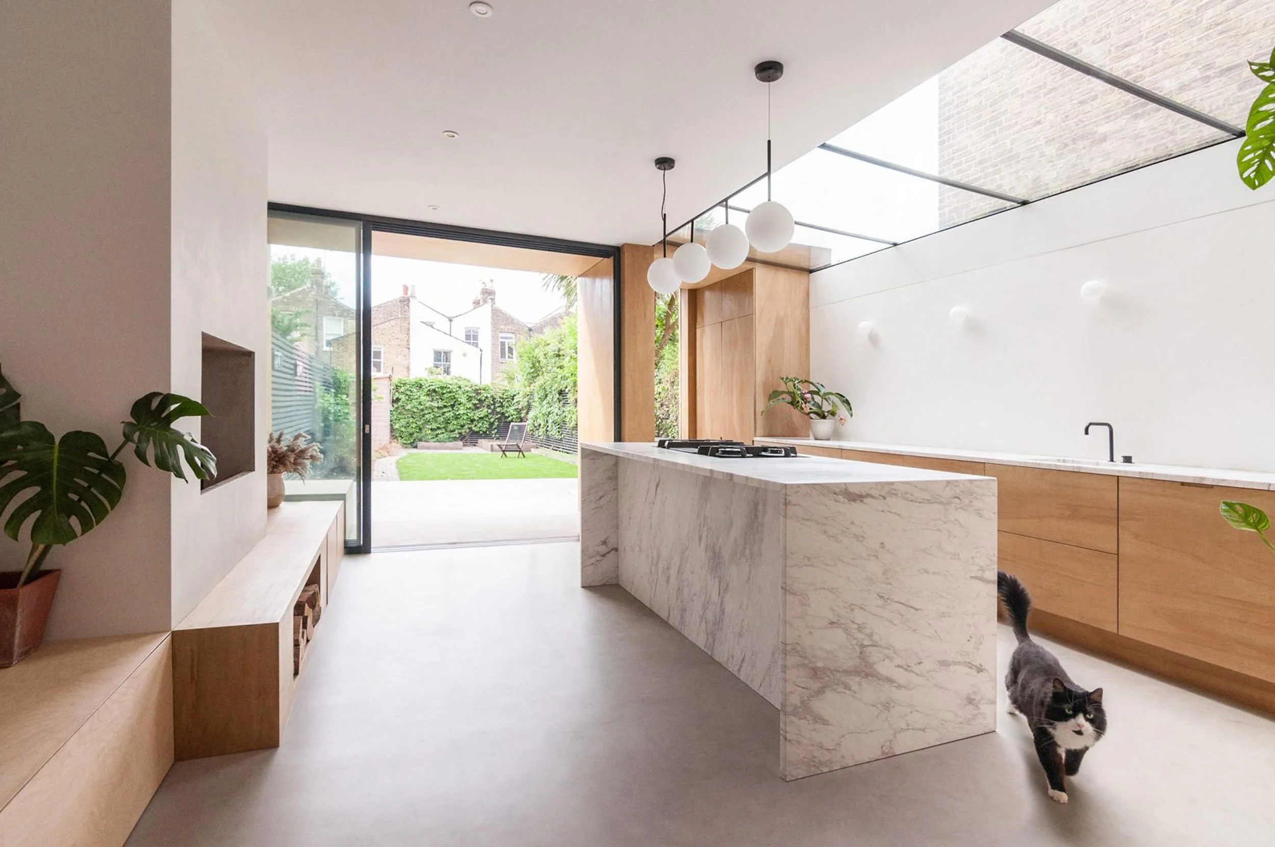 Modern kitchen with marble island, wooden cabinets, potted plants, sliding glass door opening to a backyard garden, and a black and white cat walking underneath.