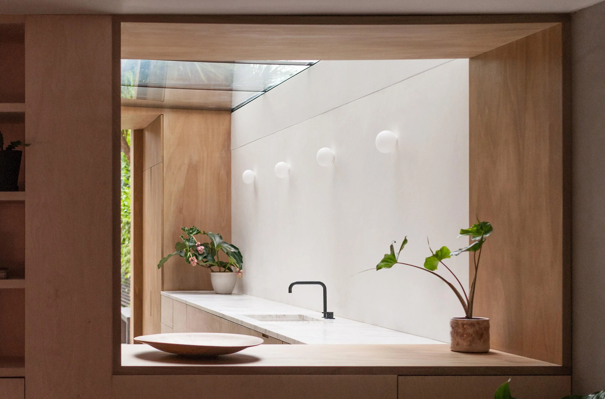 Modern kitchen with wooden frame opening, view of white counter with plants, black faucet, and wall lights