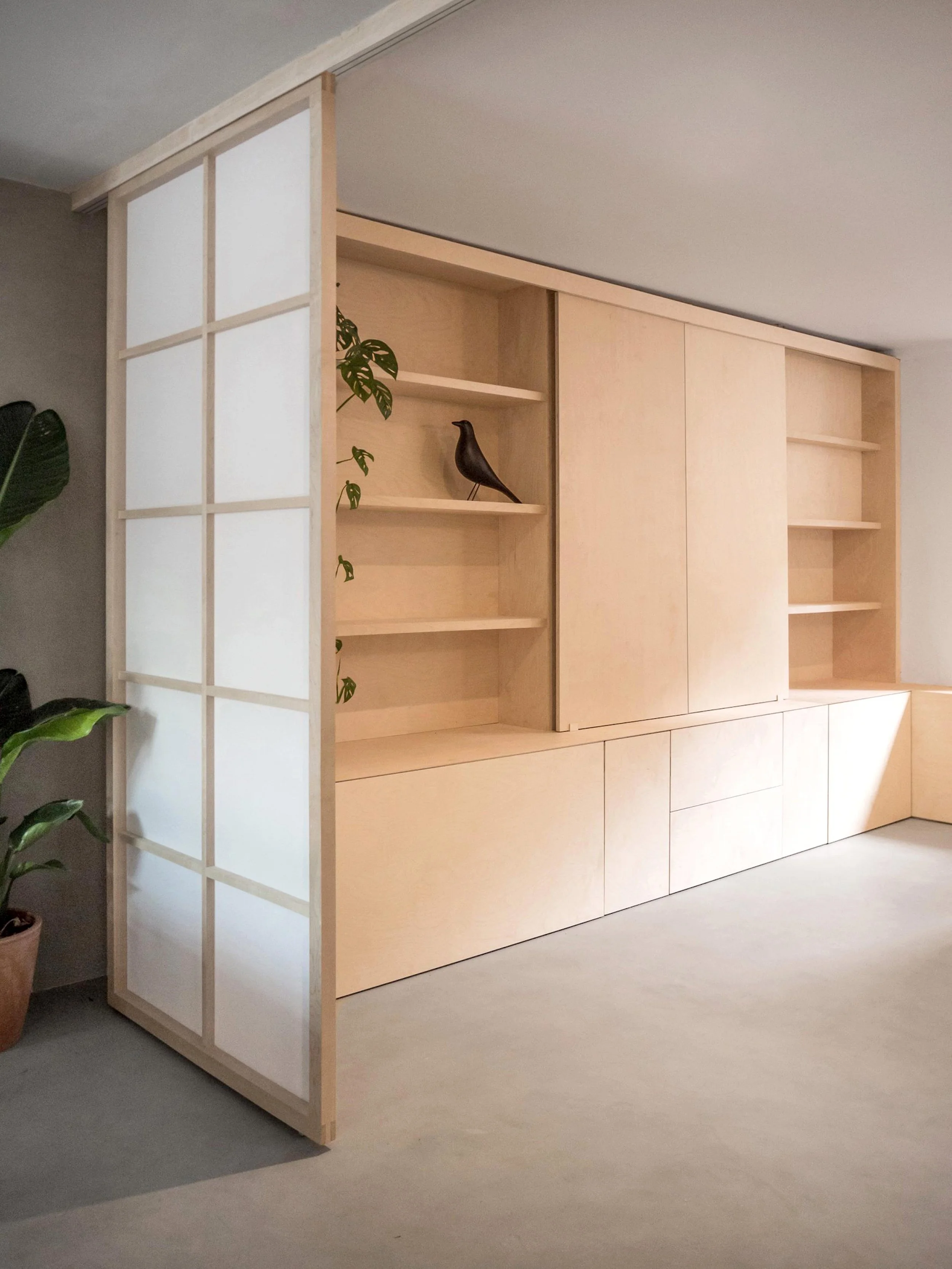 Minimalist light wood shelving unit with sliding panel doors, a black decorative bird, and green plants nearby, set in a modern interior.