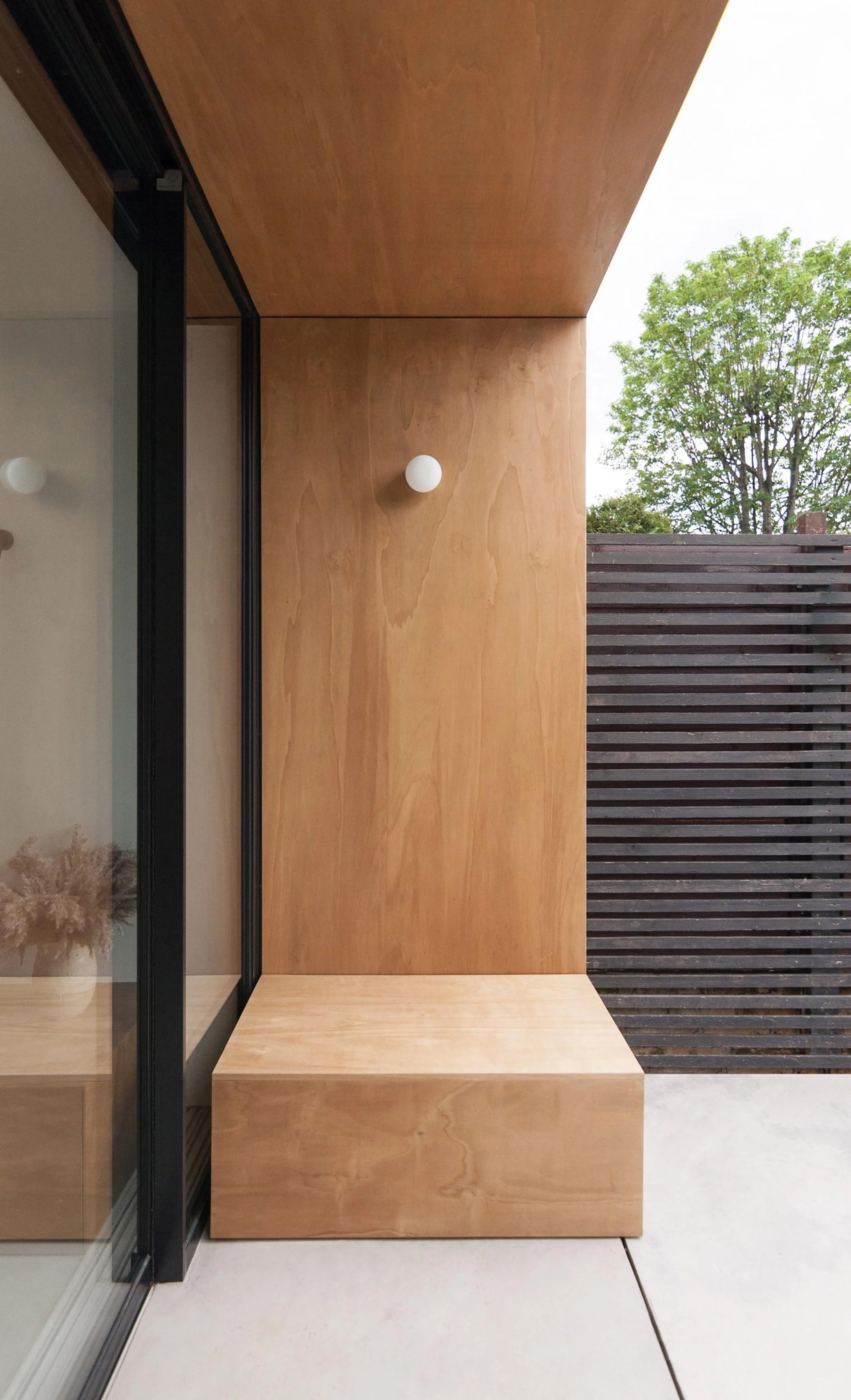 Modern outdoor patio with wooden wall, bench, and a small white spherical wall light, adjacent to a glass sliding door, with a privacy wooden fence and green tree in the background.