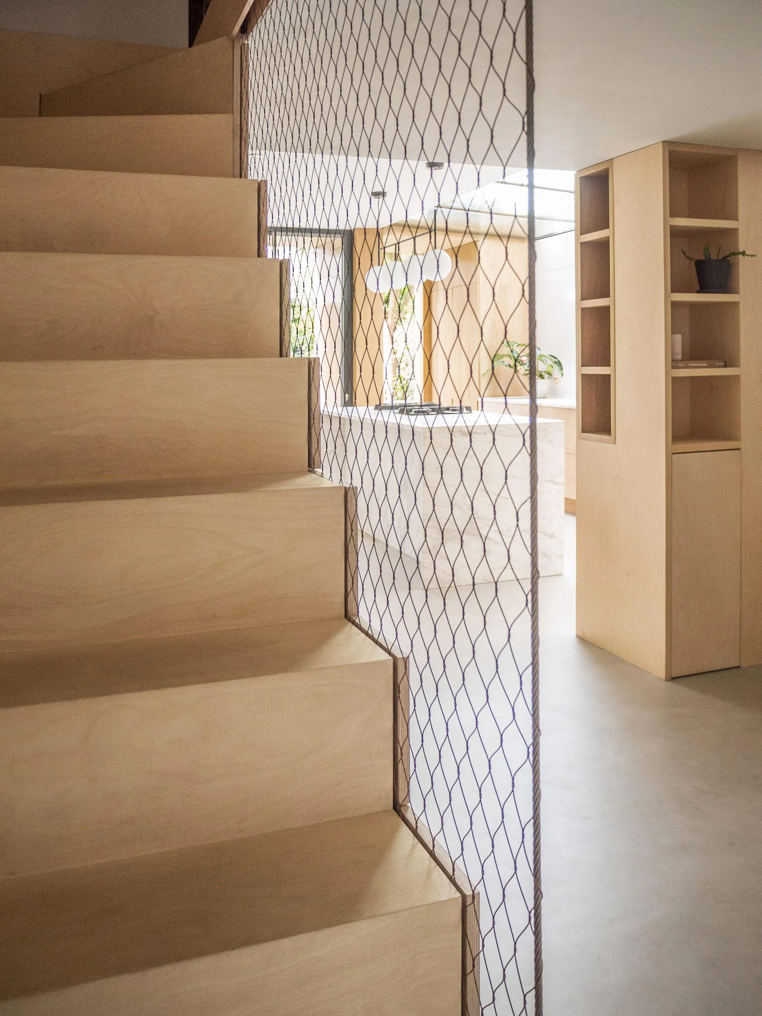 Interior view of a modern home with light wooden stairs, a mesh partition, and a kitchen area with a white island, potted plants, and wooden cabinetry.