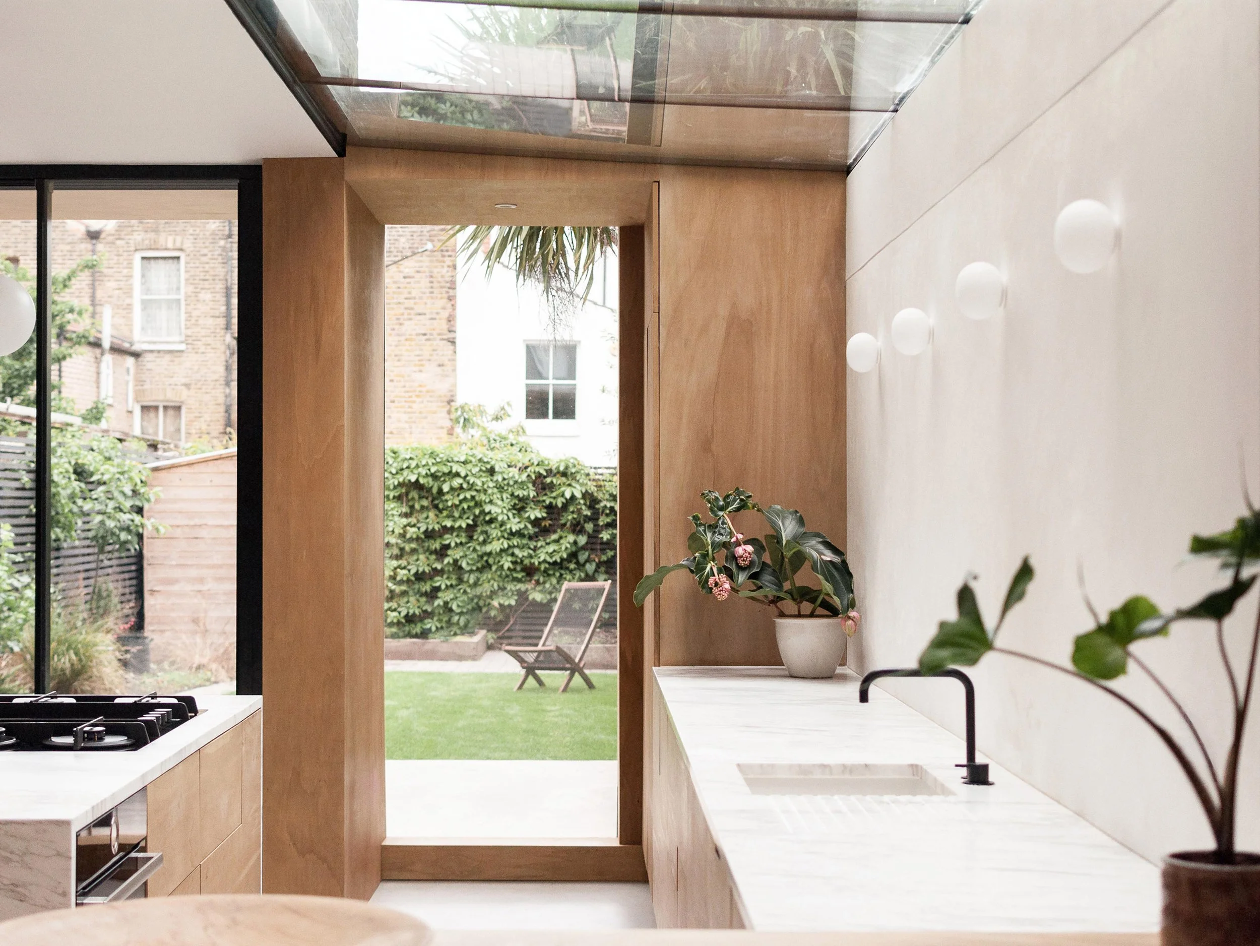 Modern kitchen with white countertops, black sink faucet, potted plant, and view of garden outside through door and window.