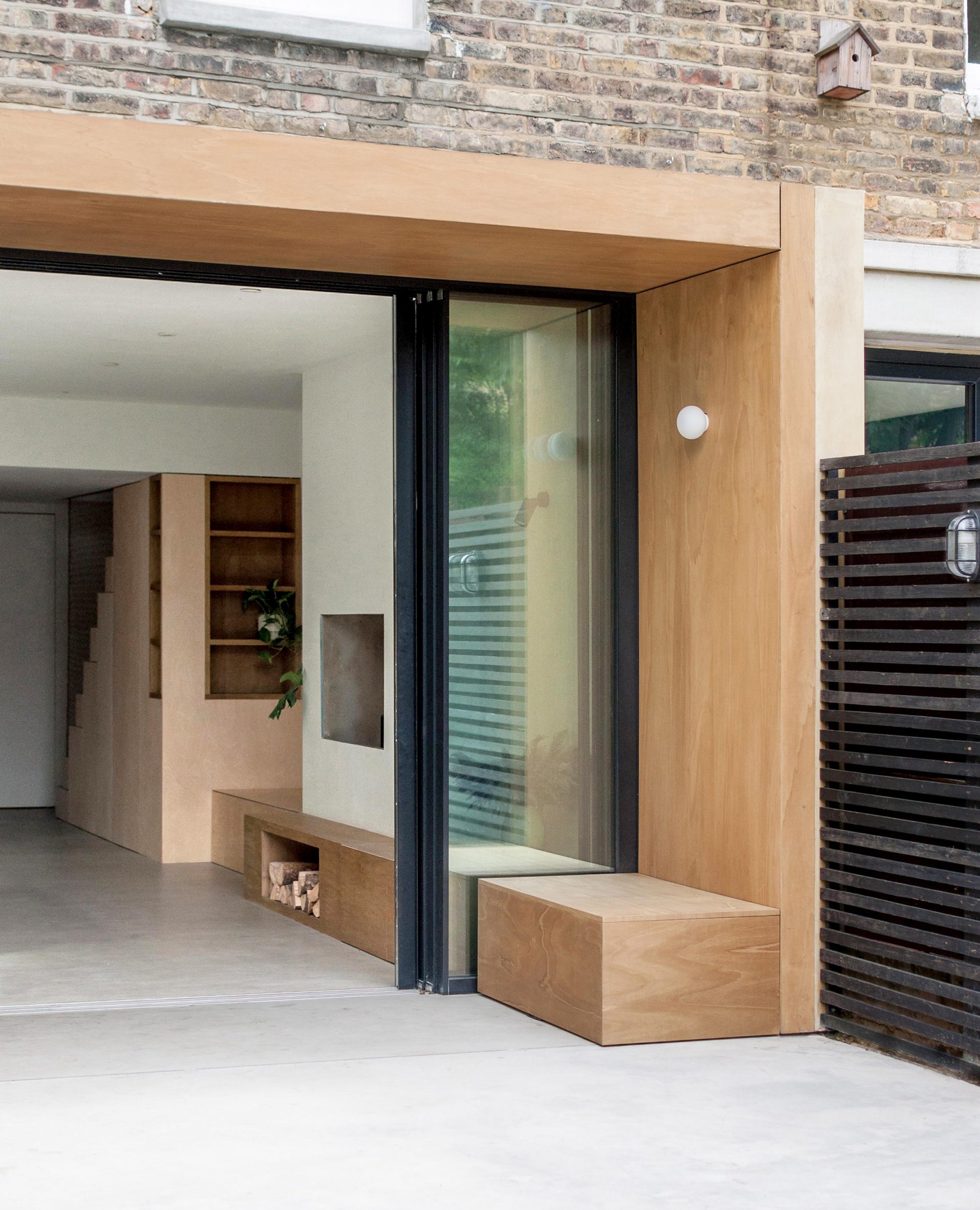 Modern house entrance with large glass door, wood accents, a built-in bench, a birdhouse on the brick wall, and a black slatted fence.